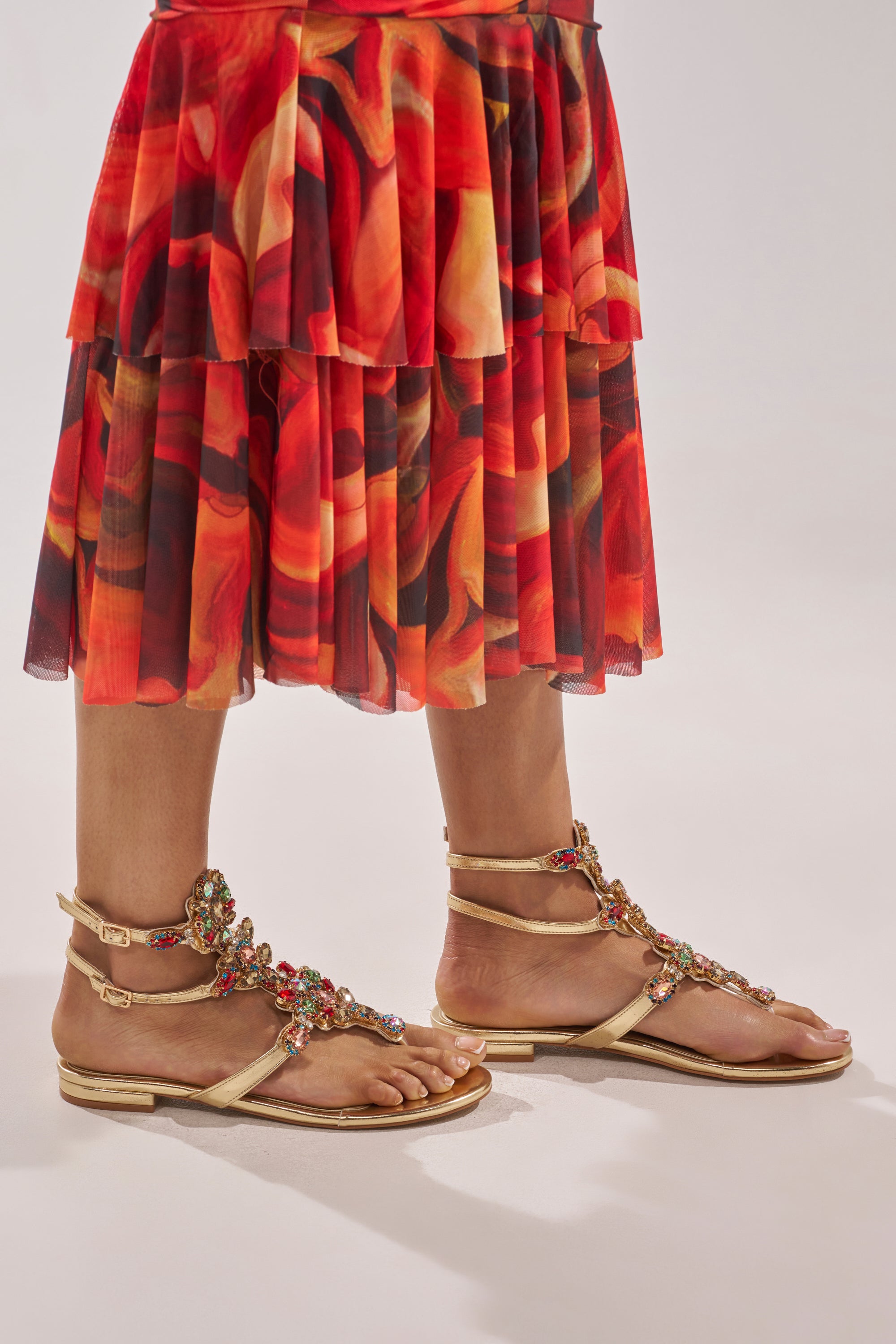 A person stands on a light background wearing a colorful, ruffled red and orange skirt and the AZALEA WANG OLISA GOLD RHINESTONE EMBELLISHED FLAT SANDAL adorned with red and green details. 2
