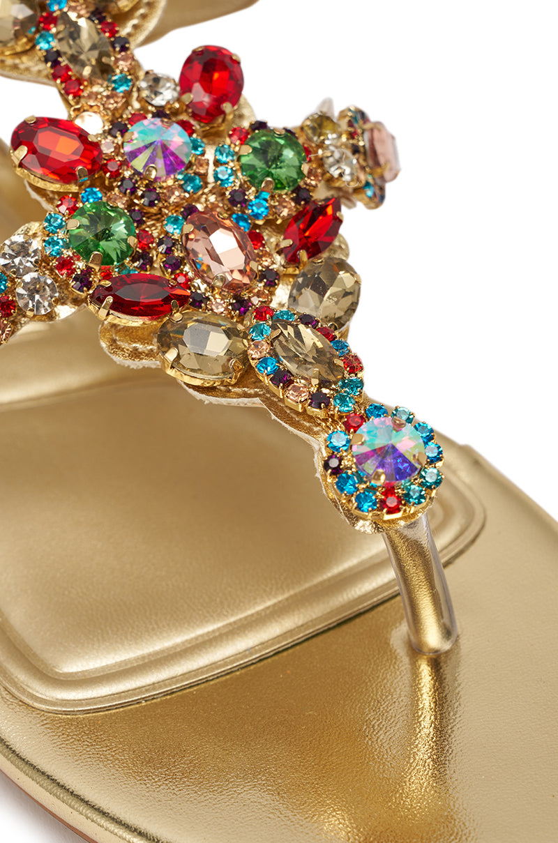 A close-up of the AZALEA WANG OLISA Gold Rhinestone Embellished Flat Sandal, showcasing an ornate T-strap with red, green, blue, and clear gemstones in intricate crystal patterns. 7