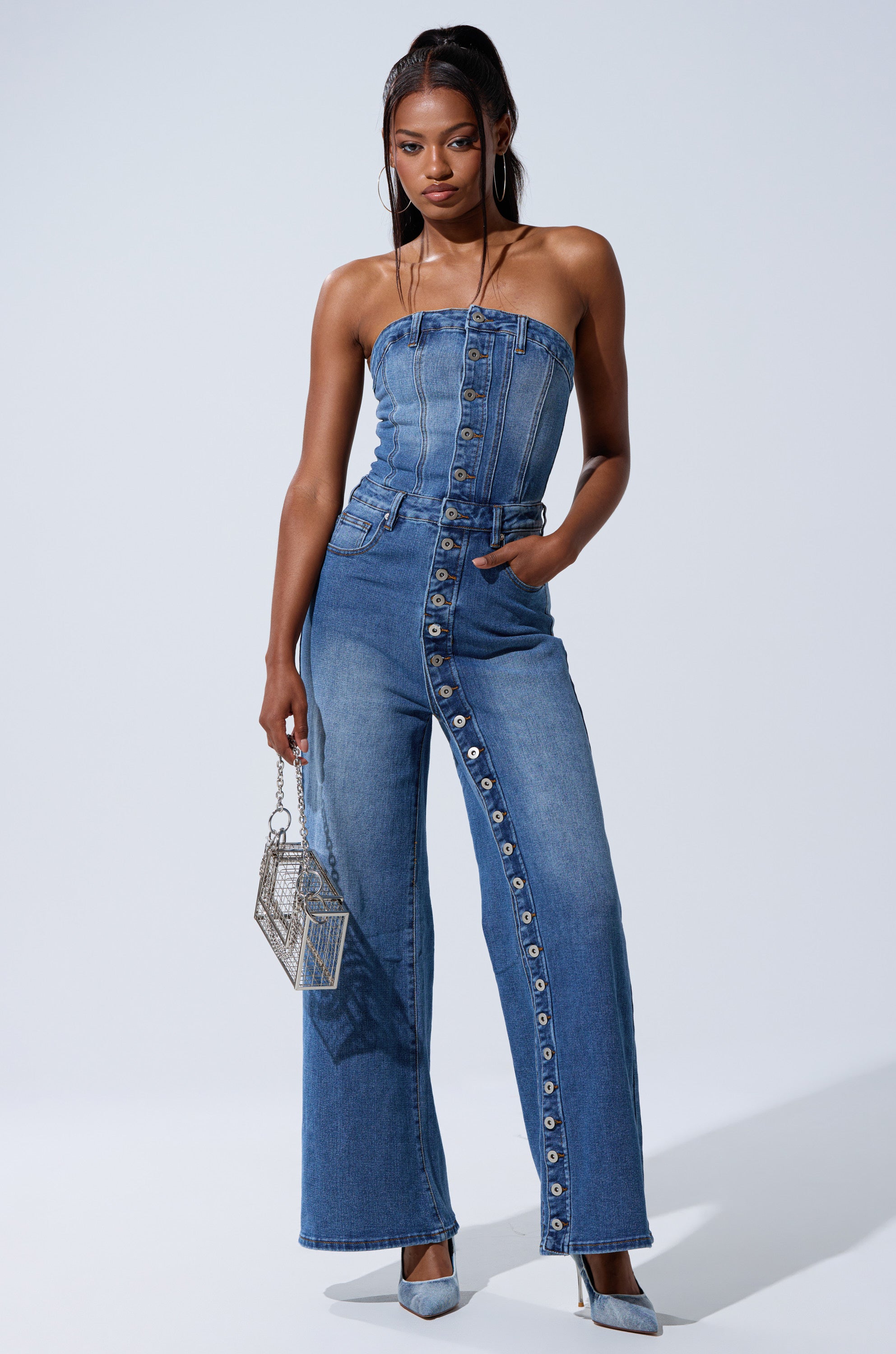 A woman models the SUMMER SONG DENIM JUMPSUIT, a strapless piece with wide legs and metal buttons, one hand in her pocket. She pairs it with blue heels and a silver handbag, posing against a plain light background.