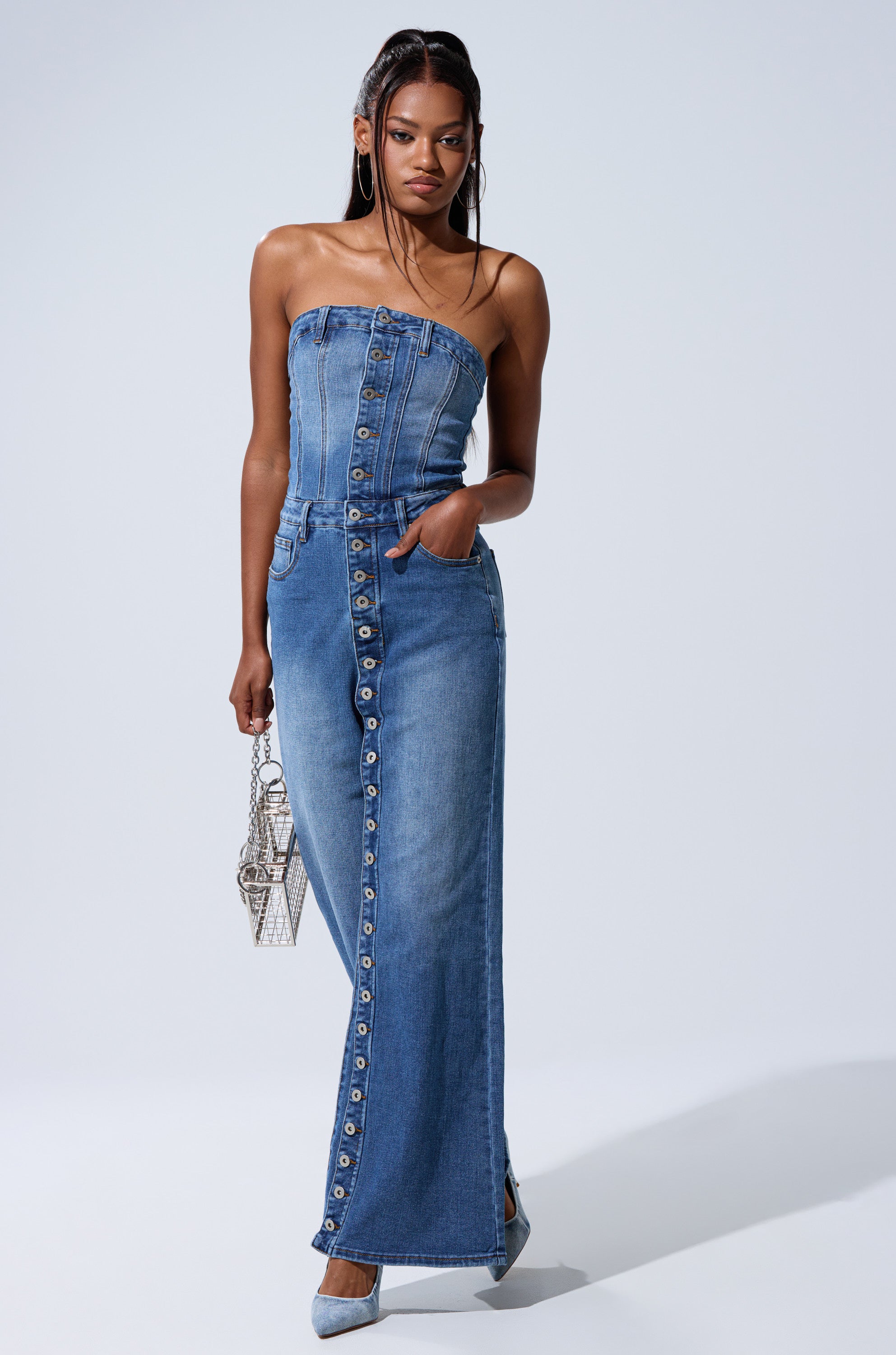 A woman models the SUMMER SONG DENIM JUMPSUIT, a strapless style with one hand in her pocket and a small silver purse. She accessorizes with hoop earrings and pointed heels, set against a light gray background.