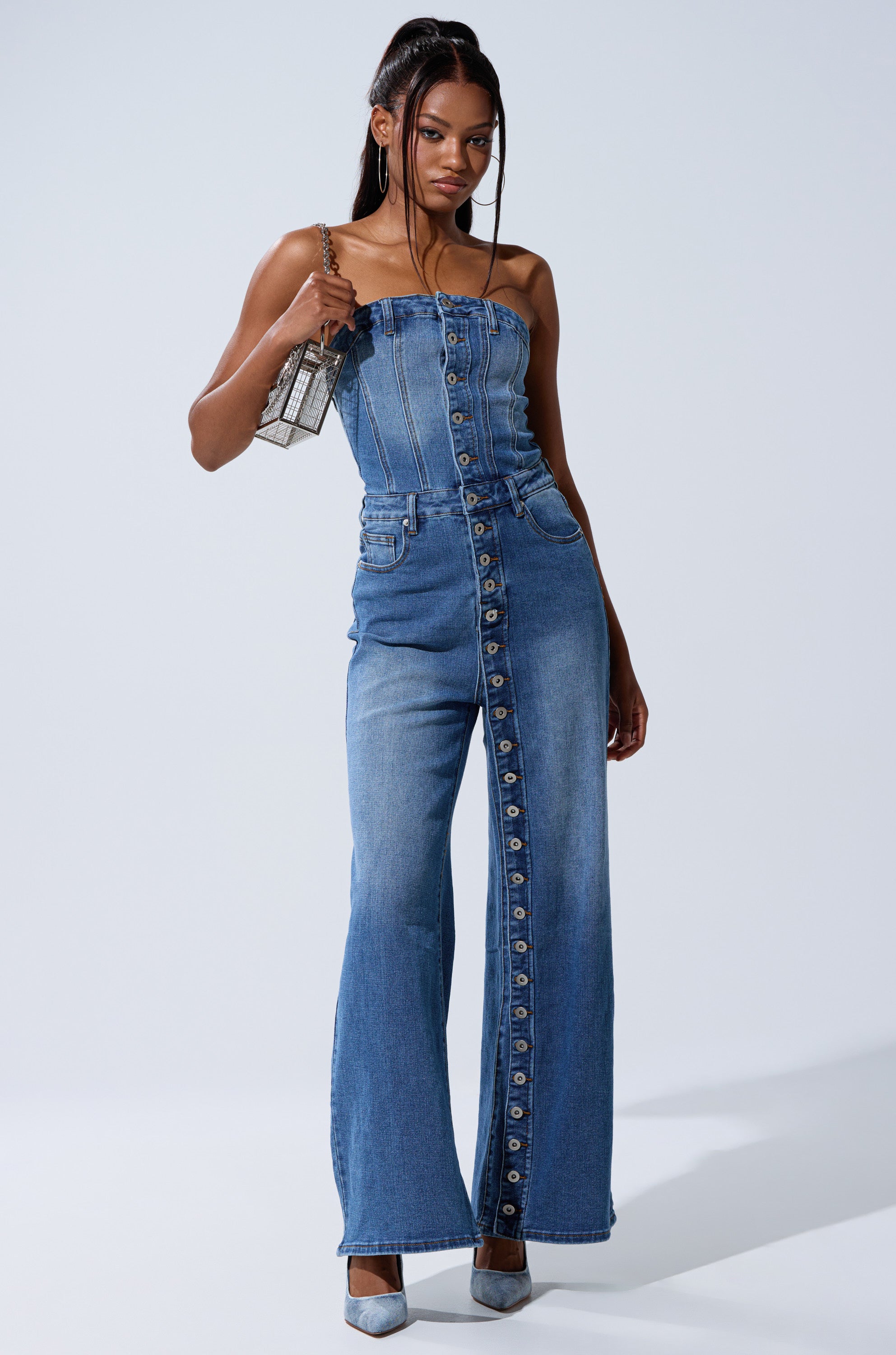 A woman models the SUMMER SONG DENIM JUMPSUIT, featuring a strapless design and wide, flared legs. She accessorizes with a silver handbag, hoop earrings, silver heels, and wears her hair in high pigtails against a light background.