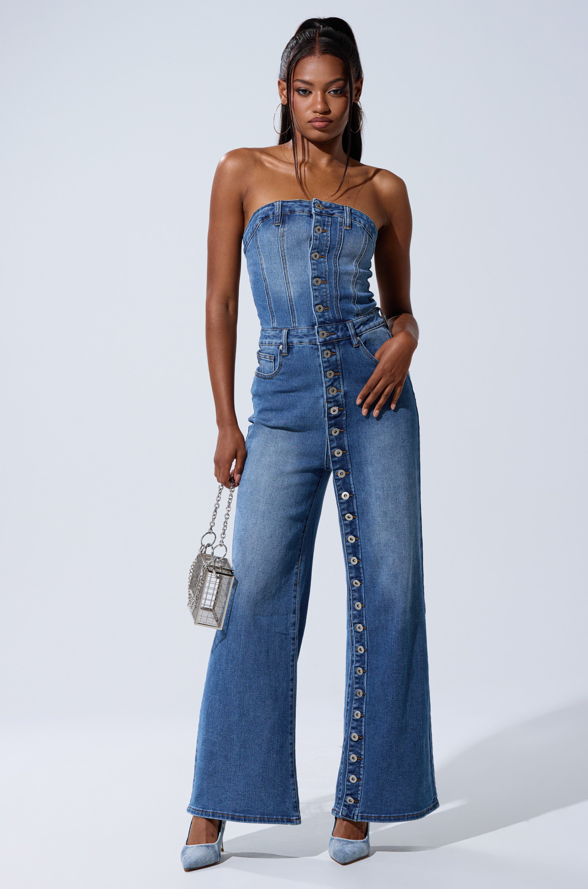 A woman poses confidently in the SUMMER SONG DENIM JUMPSUIT, featuring a strapless top and wide-leg pants. She pairs it with light blue heels and a small silver handbag against a plain light background.