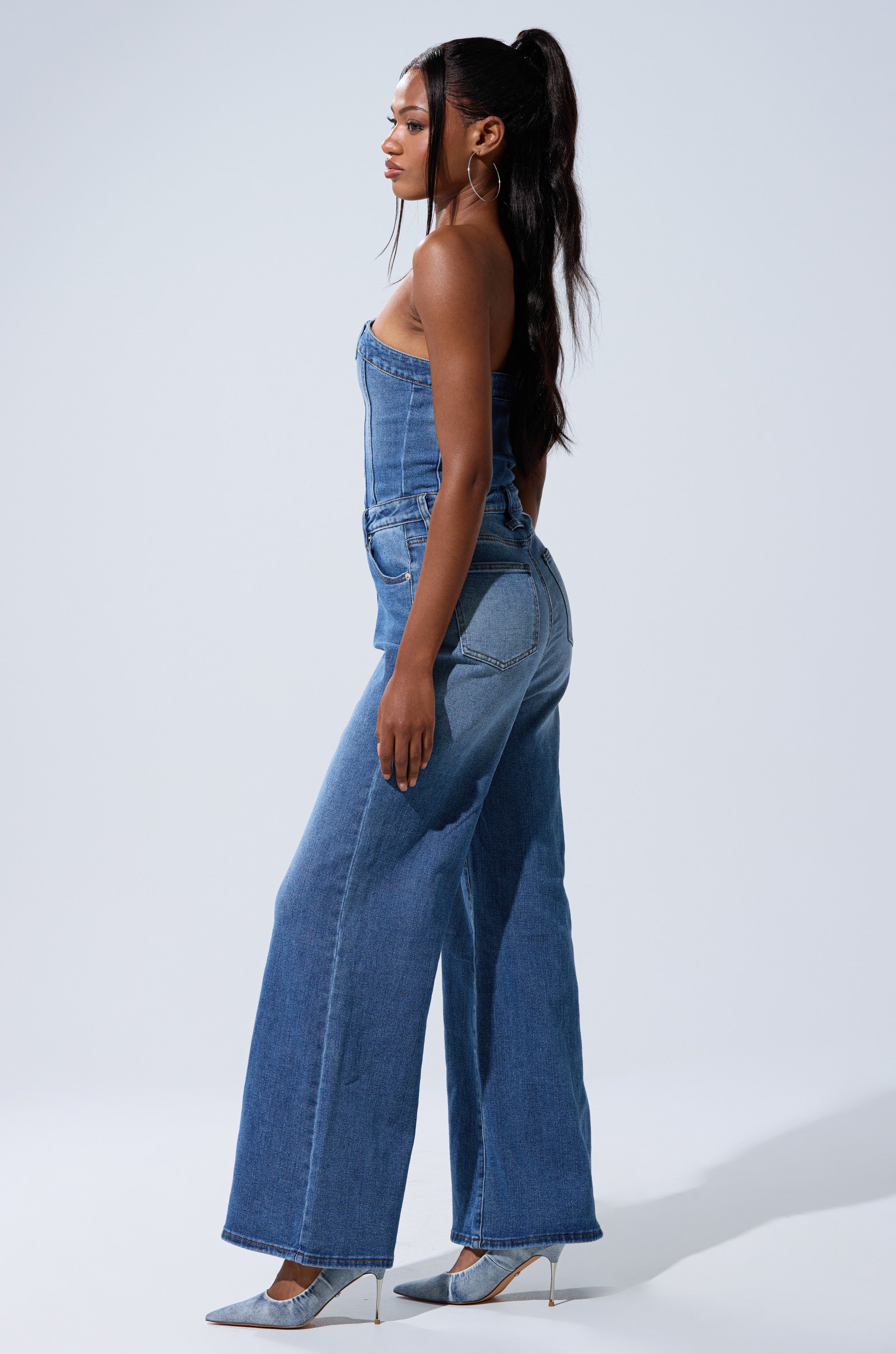 A woman with long dark hair in a high ponytail stands in profile against a light background, wearing the chic SUMMER SONG DENIM JUMPSUIT with pointed-toe silver heels.
