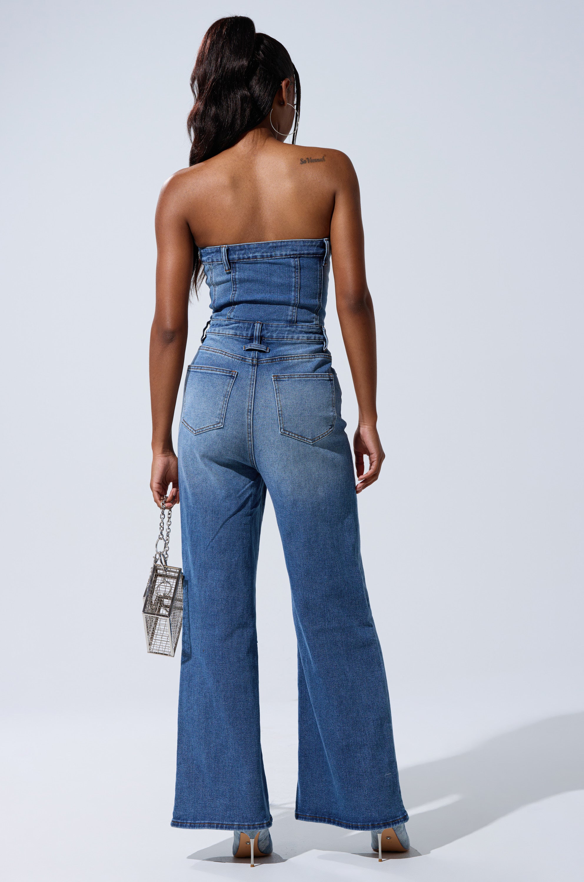 A woman faces away in the SUMMER SONG DENIM JUMPSUIT—strapless, with flared legs and faded details. She carries a clear handbag, wears heels, and has her hair in a high ponytail against a plain light background.