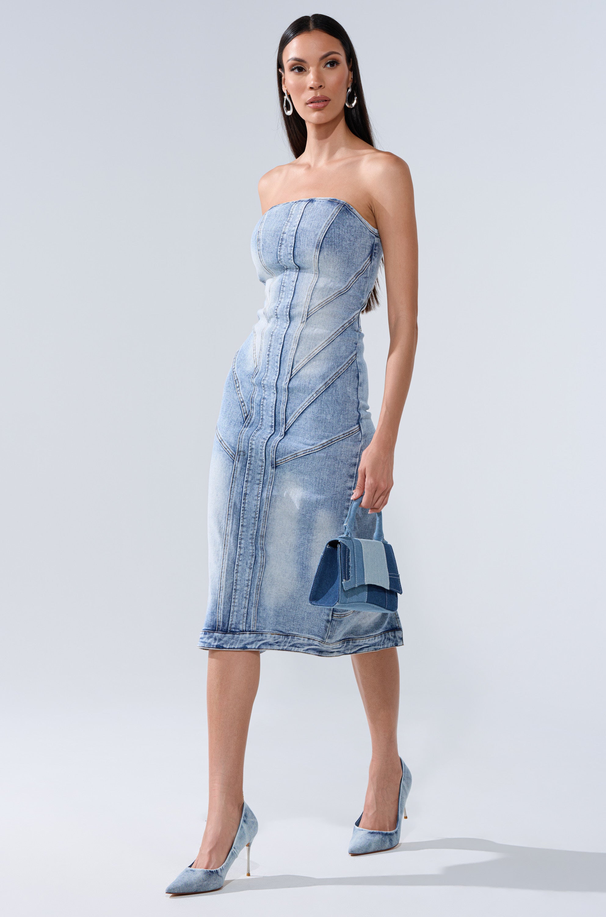 A woman wearing the BROOKLYN BADDIE DENIM MIDI DRESS, paired with light blue heels and a small blue handbag, stands against a plain light background.