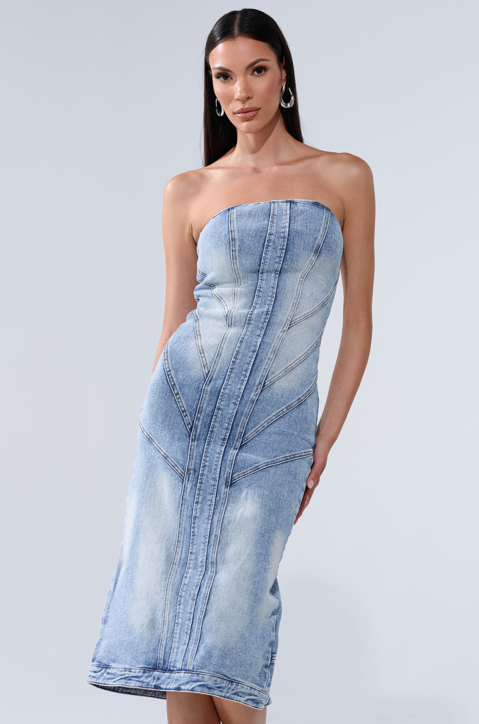 A woman in the BROOKLYN BADDIE DENIM MIDI DRESS—a strapless, form-fitting light wash midi with visible stitching—stands against a plain light gray background. She has long straight hair and wears hoop earrings.