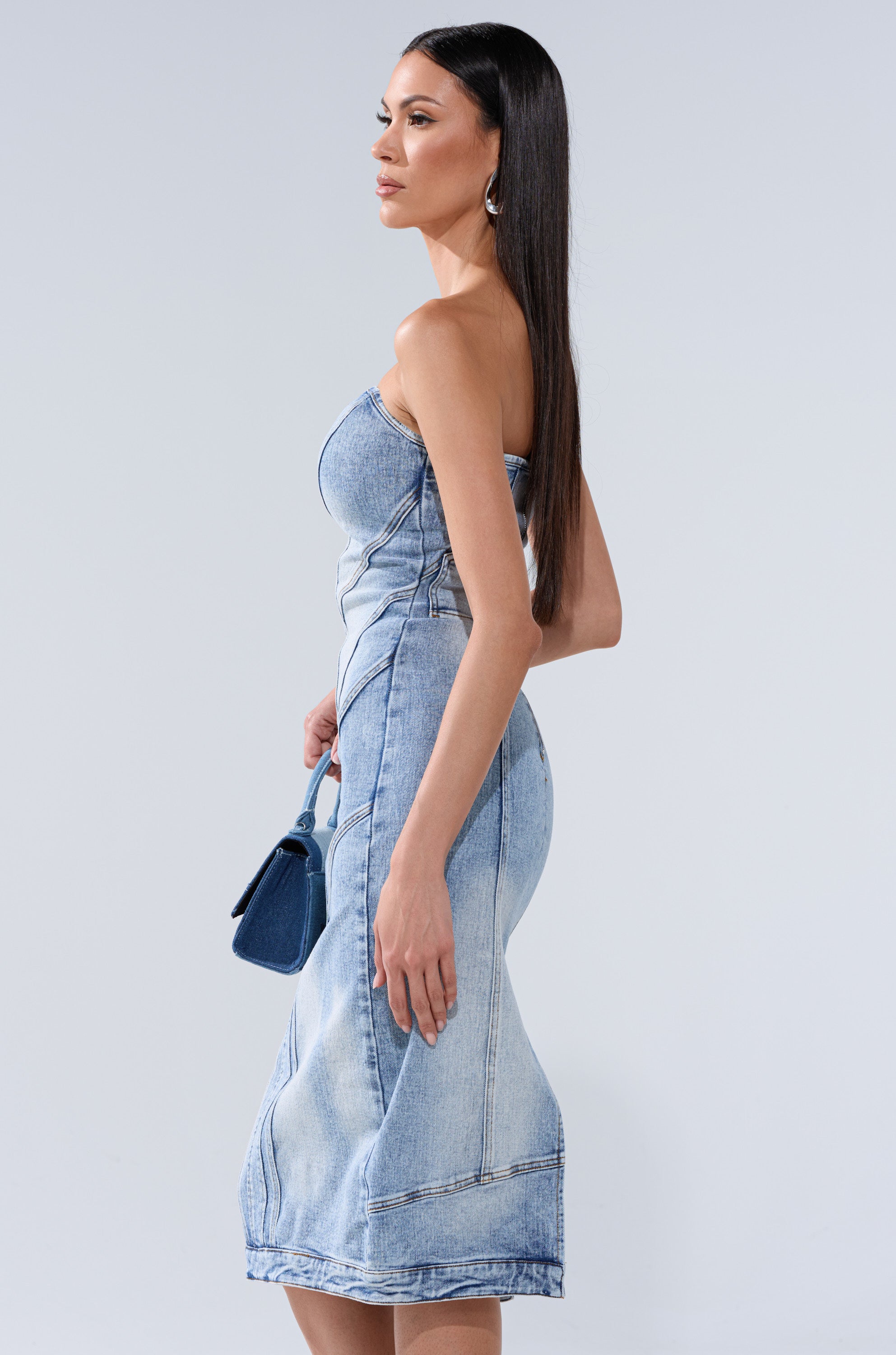 A woman with long straight hair wears the BROOKLYN BADDIE DENIM MIDI DRESS and holds a small blue handbag, standing in profile against a plain light gray background.