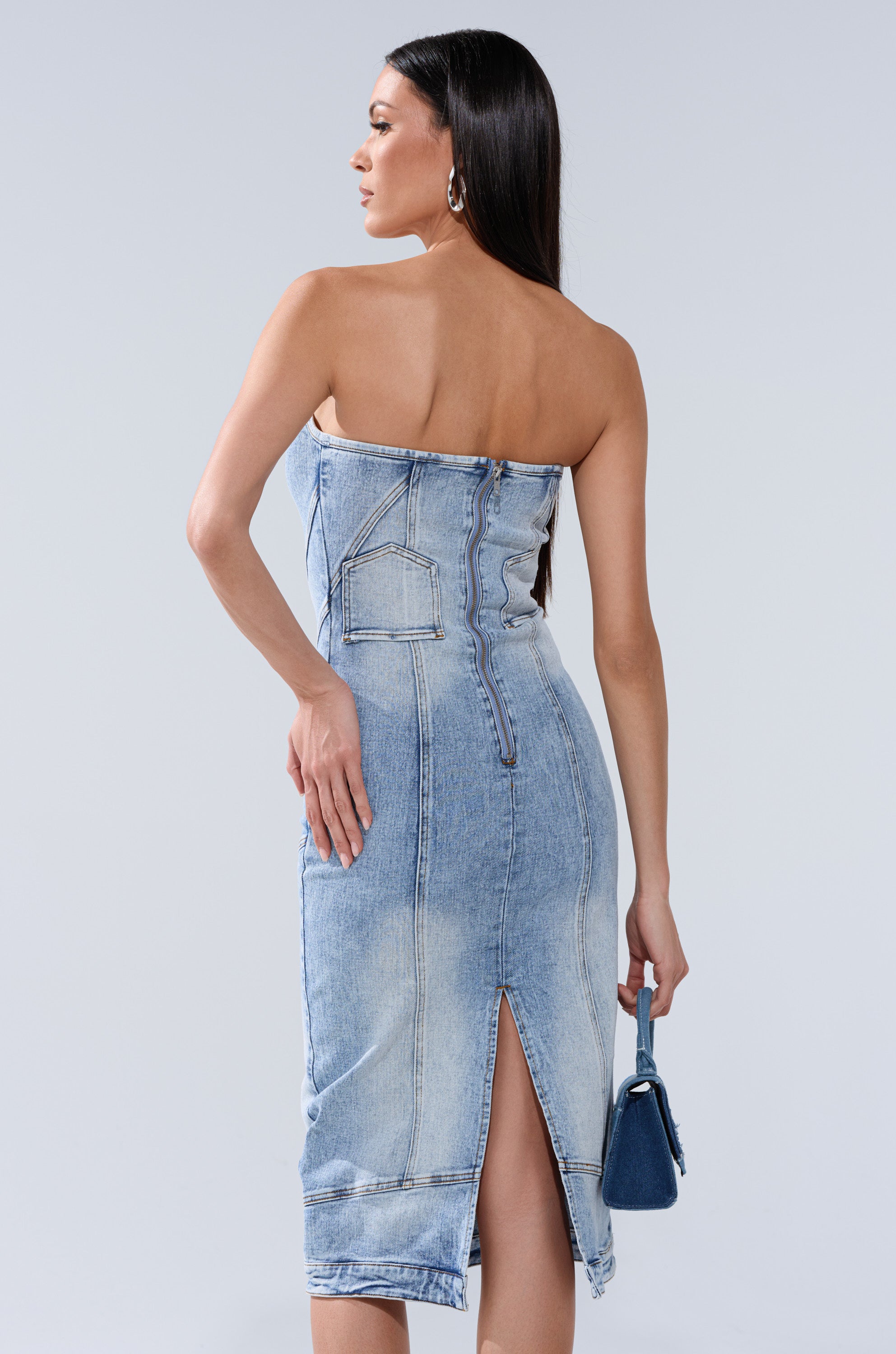 A woman with long dark hair wears the BROOKLYN BADDIE DENIM MIDI DRESS, a light blue strapless midi with visible stitching, back slit, and small back pocket, as she stands facing away holding a small blue handbag.
