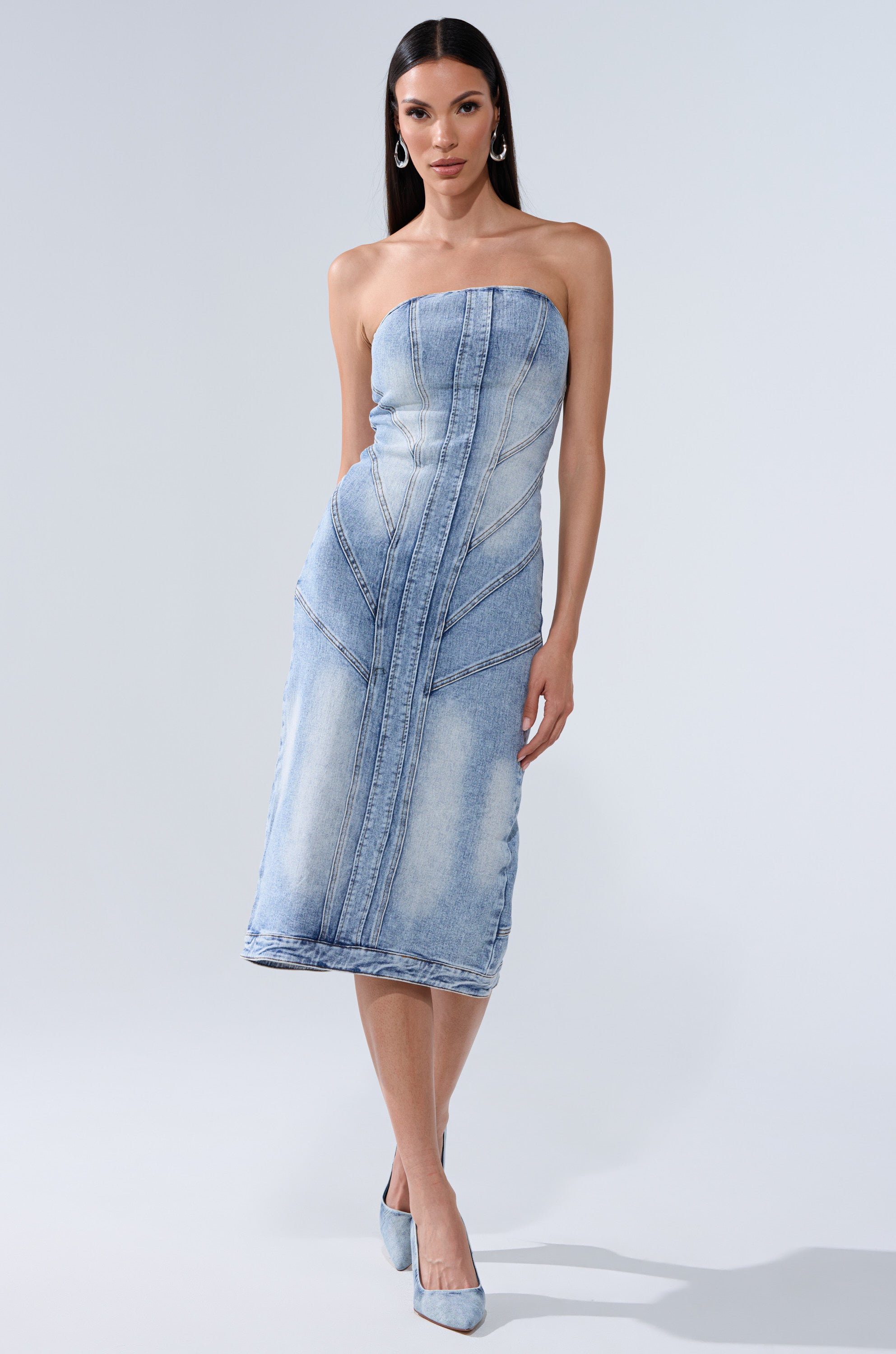 A woman models the BROOKLYN BADDIE DENIM MIDI DRESS, a strapless, light-wash denim midi with vertical seams. She completes the look with matching denim heels, hoop earrings, and sleek hair against a plain background.