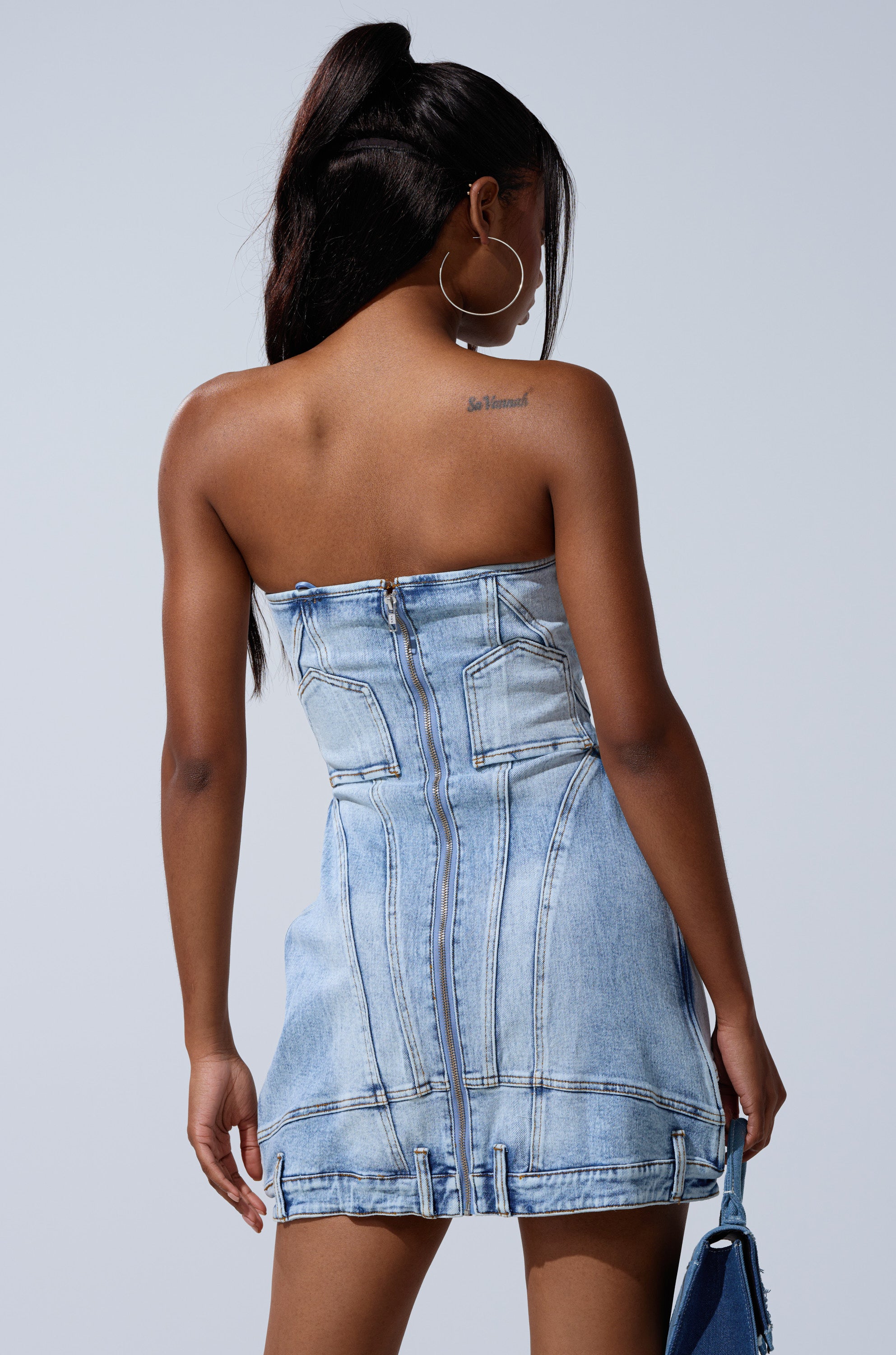 A woman with long dark hair in a high ponytail wears the GET LIKE ME DENIM MINI DRESS, strapless and light-wash, facing away to reveal a tattoo on her right shoulder blade while holding a blue bag by her side.