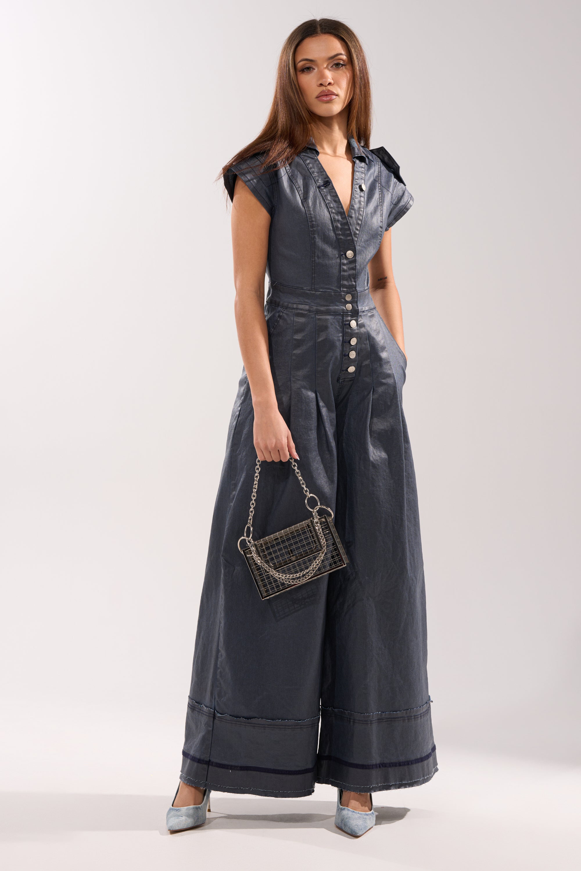 A woman wears the RONNIE DENIM JUMPSUIT, featuring wide legs, a dark wash, short sleeves, and front buttons. She accessorizes with a chain-strap purse and light blue heels. Her long brown hair is straightened. 0
