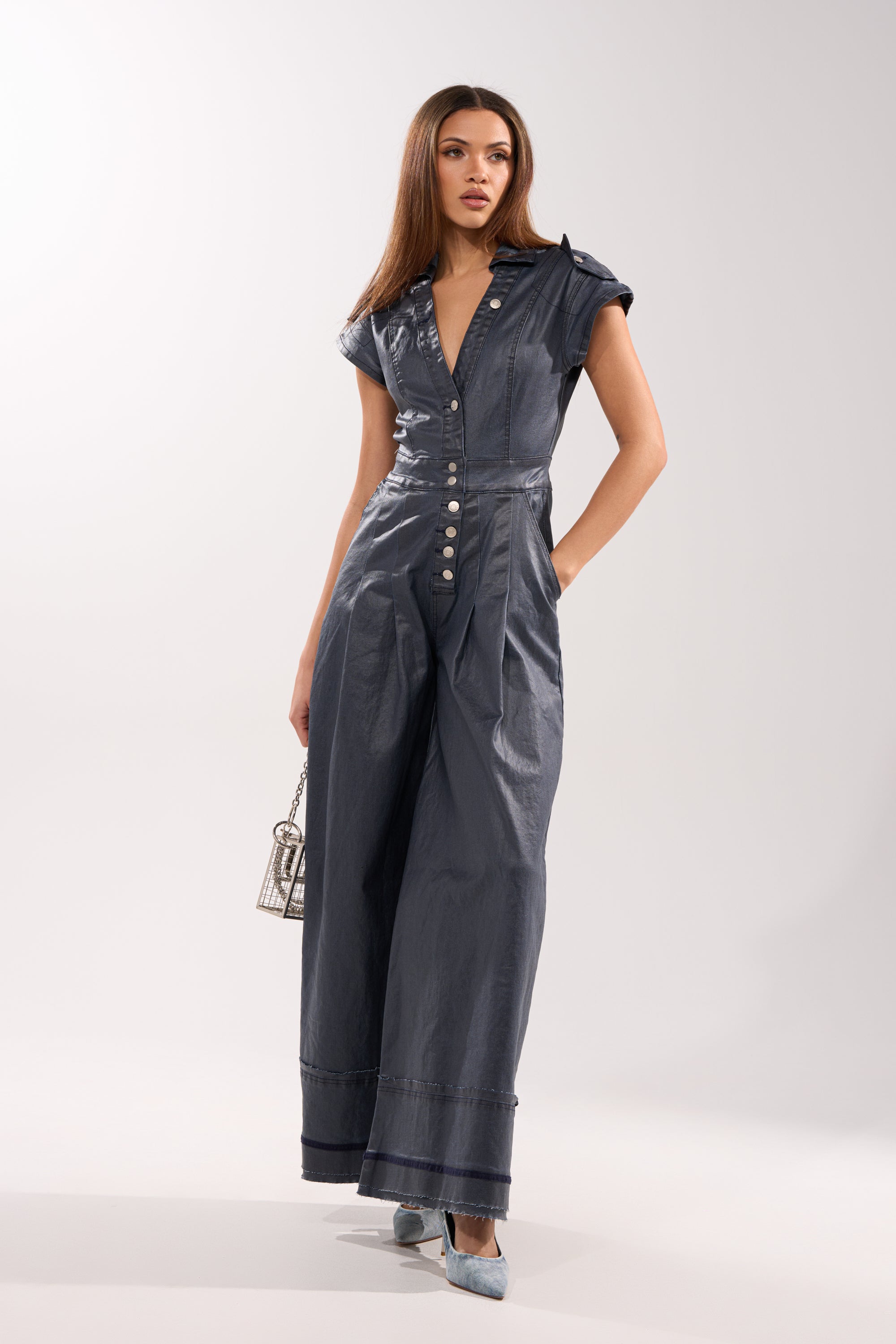 A woman wears the RONNIE DENIM JUMPSUIT, featuring a shiny dark wash, short sleeves, wide legs, and prominent front buttons. She accessorizes with a small purse and light blue heels against a plain light background. 1