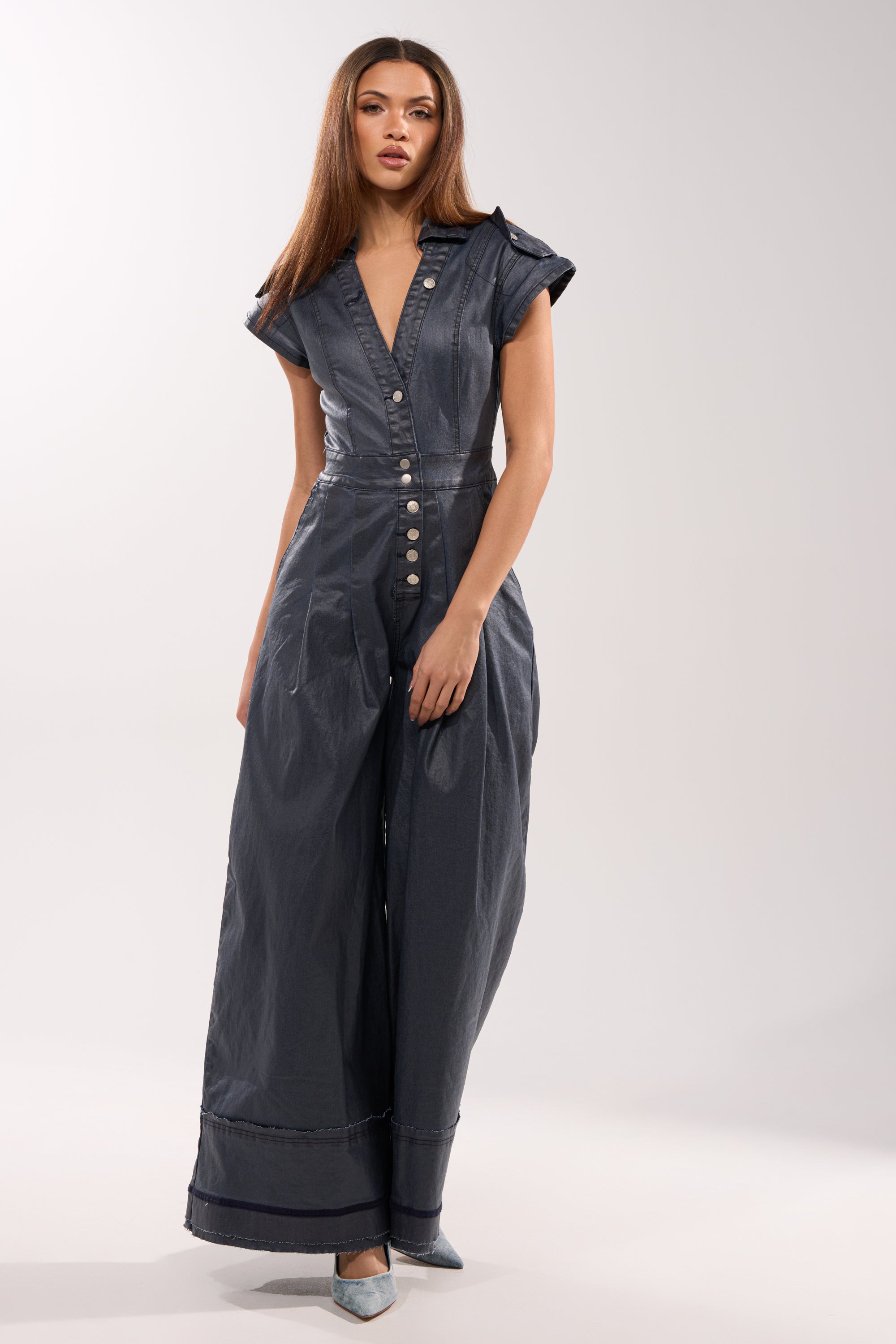 A woman wears the RONNIE DENIM JUMPSUIT, a short-sleeved, button-down, wide-leg style in stretch denim with a deep V-neck, paired with light blue pointed-toe heels, against a simple light background. 2