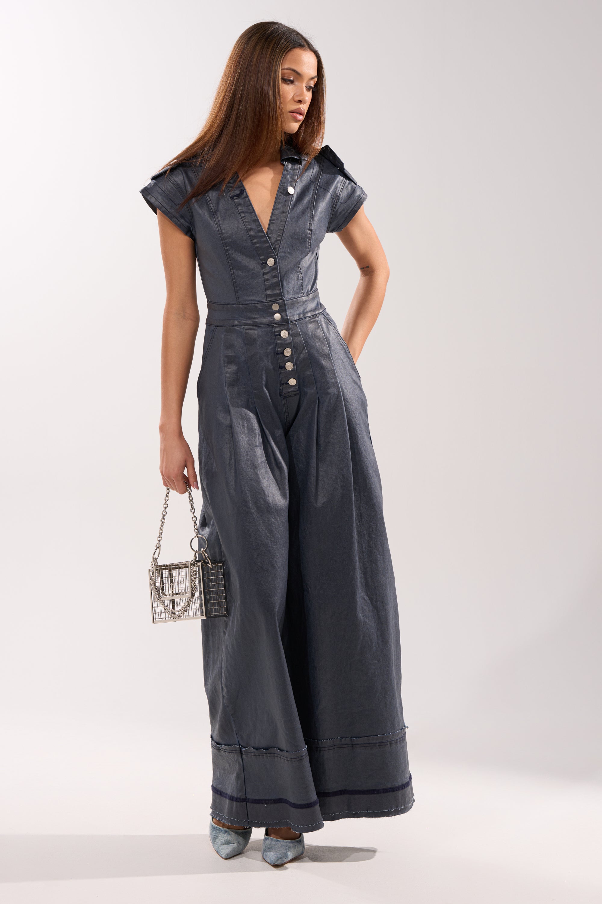 A woman models the RONNIE DENIM JUMPSUIT, featuring wide-leg, dark blue denim with short sleeves and front buttons. She accessorizes with a metallic handbag, pointed-toe heels, and wears her straight brown hair loose. 3