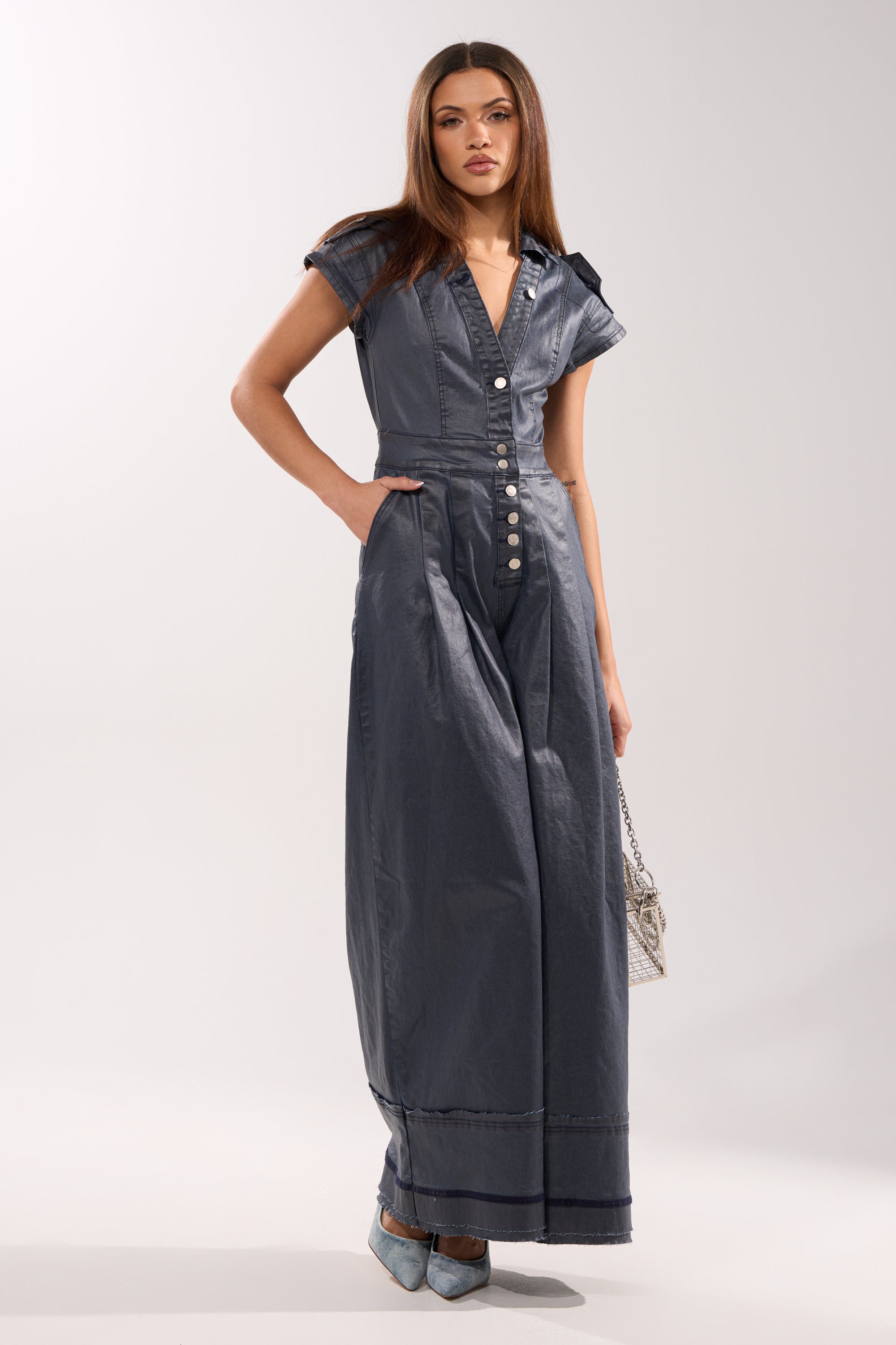 A woman with long brown hair and a neutral expression wears the RONNIE DENIM JUMPSUIT—wide-leg, short-sleeve, button-front—styled with blue pointed-toe heels and a small patterned handbag against a plain background. 4
