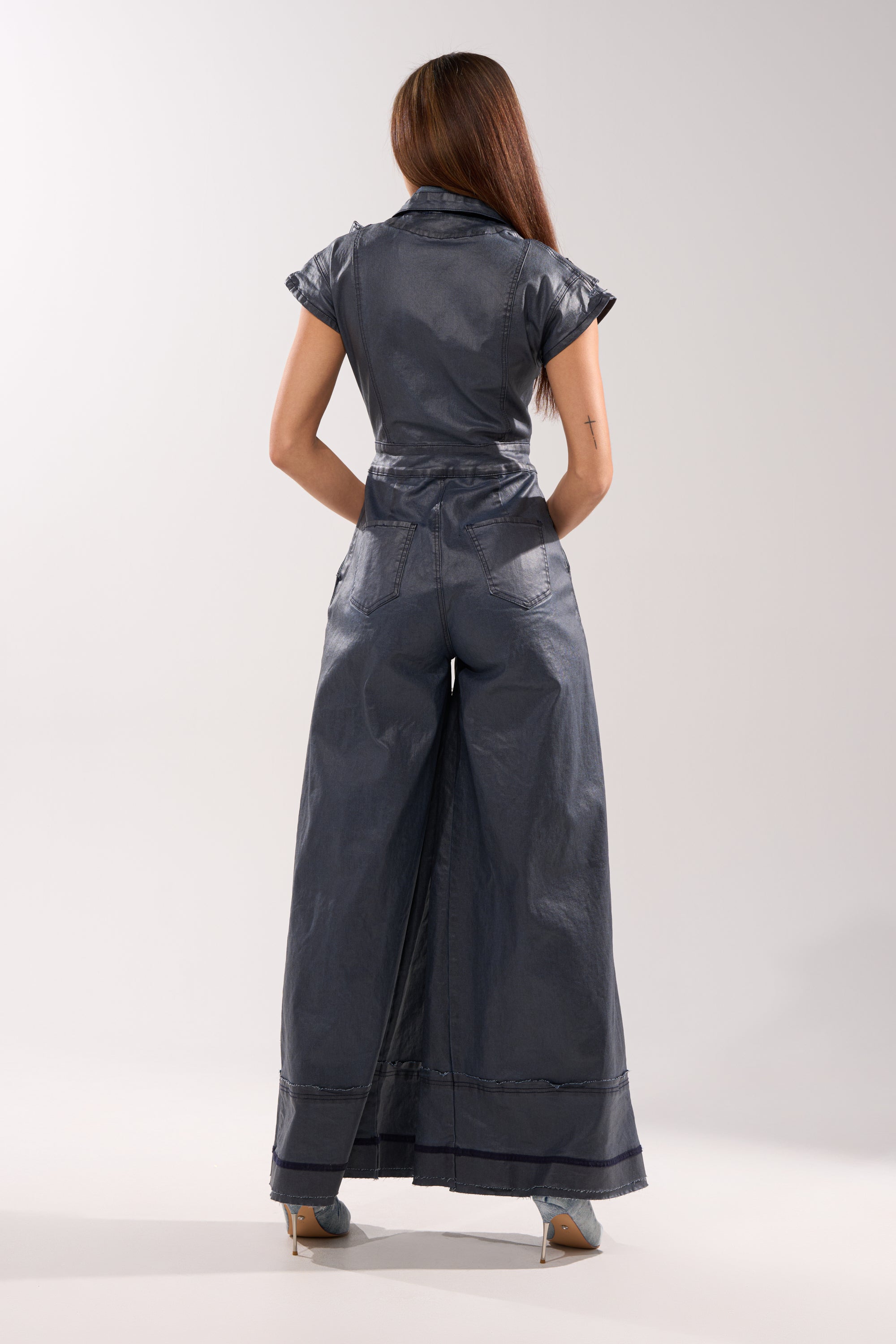 A person with long brown hair stands facing away with hands in back pockets, wearing the RONNIE DENIM JUMPSUIT—short-sleeved, belted, wide-leg, stretch denim—and heeled shoes against a plain light background. 6