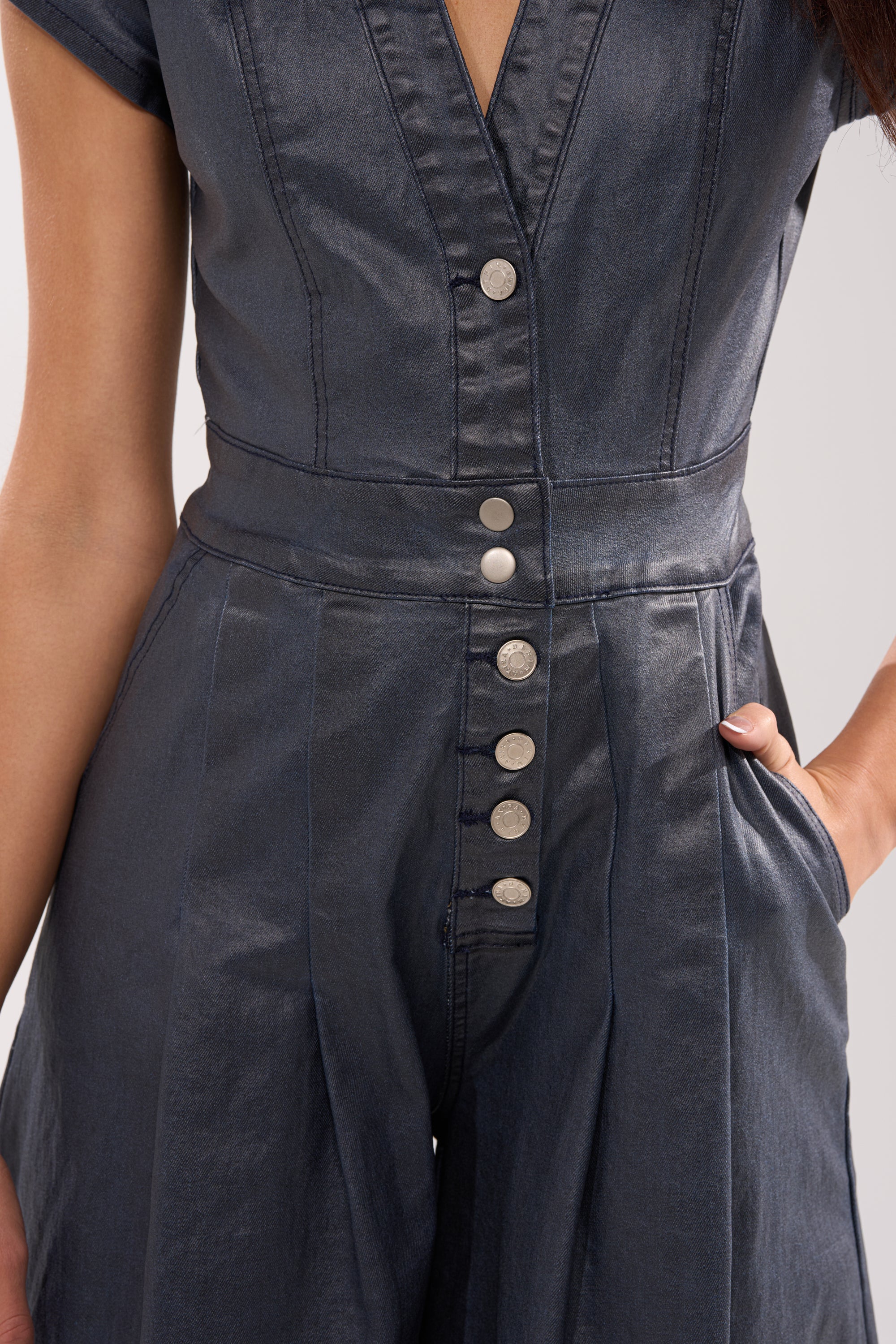 Someone wearing the RONNIE DENIM JUMPSUIT, a short-sleeve dark wash style with metallic front buttons and wide-leg cut, stands with one hand in their pocket. The jumpsuit is shown from torso to upper legs. 7