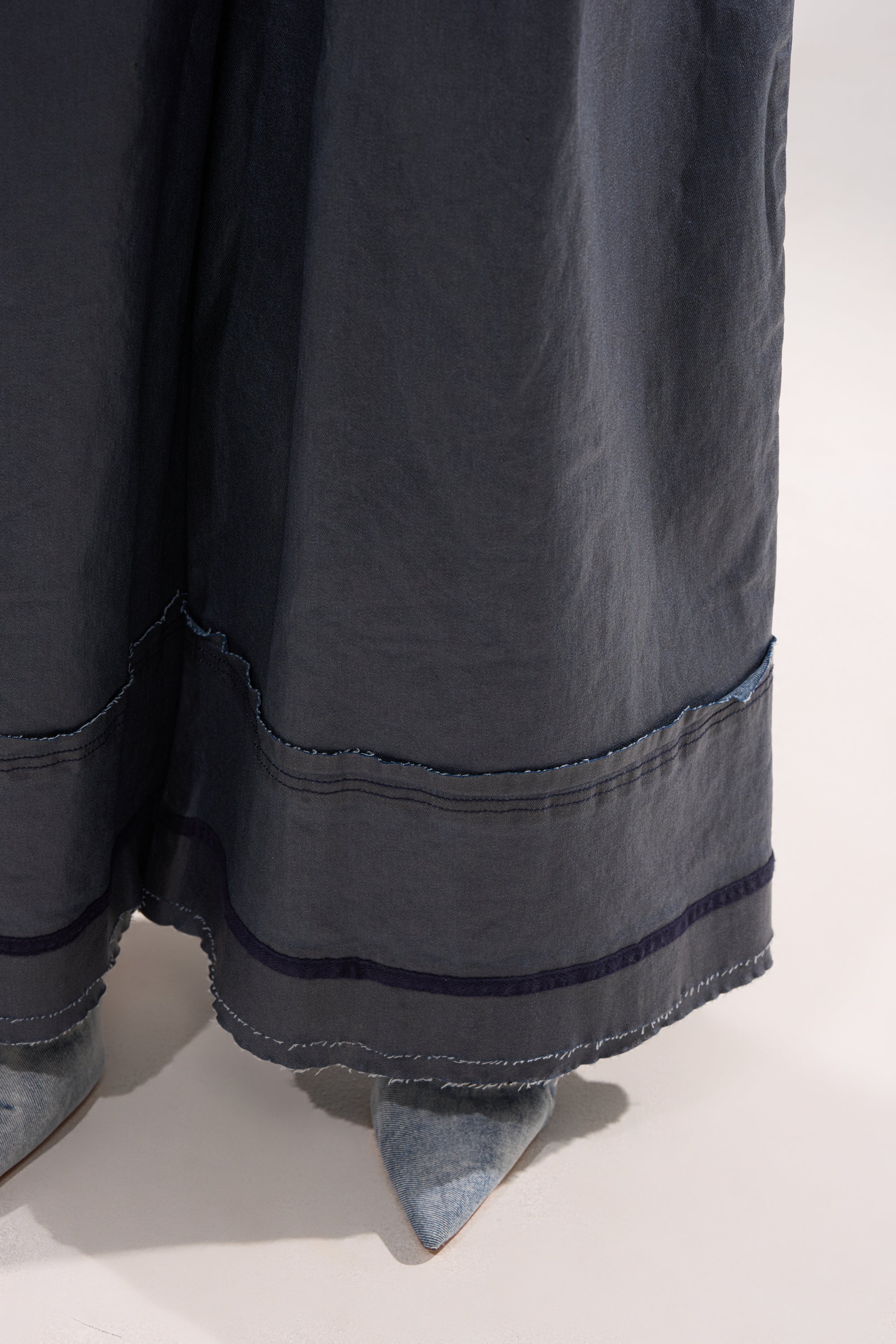 A close-up of the lower part of the RONNIE DENIM JUMPSUIT, featuring wide legs and visible stitching, paired with pointed-toe grey textured shoes on a light surface. 8