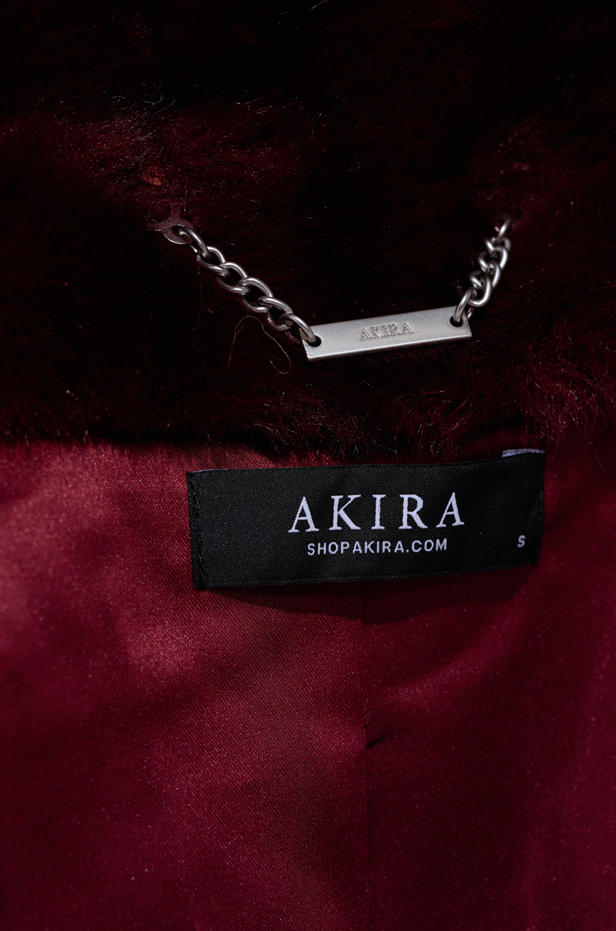 Close-up of the maroon OLYMPIA PANELED FAUX FUR shows a black AKIRA label with "SHOPAKIRA.COM," an "S" size tag, and a metal chain featuring a rectangular plate engraved "AKIRA.
