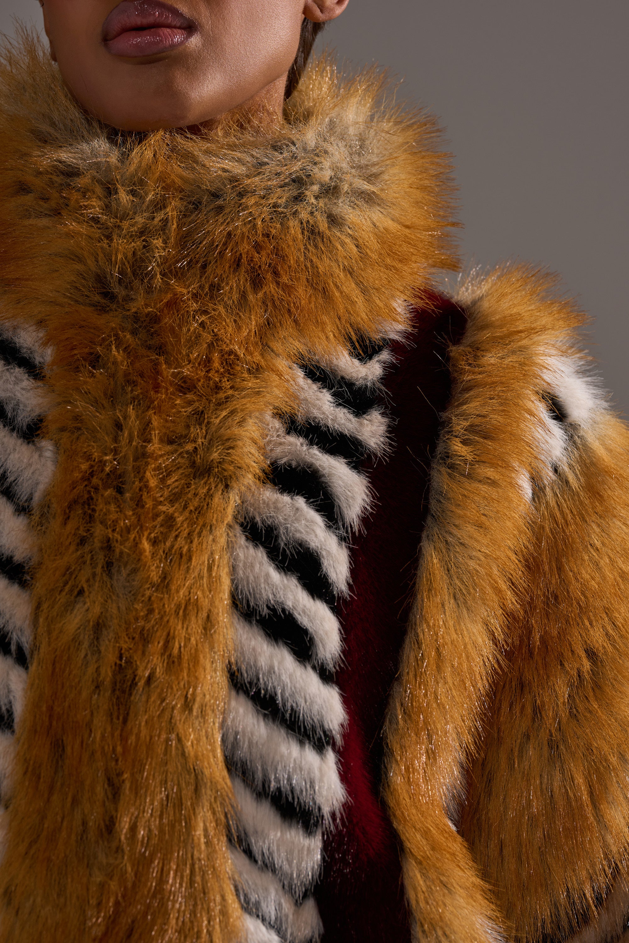 A person is shown from the shoulders up against a neutral background, wearing the OLYMPIA PANELED FAUX FUR coat with oversized panels in orange, black, white, and maroon stripes. Their lips and part of their face are visible.