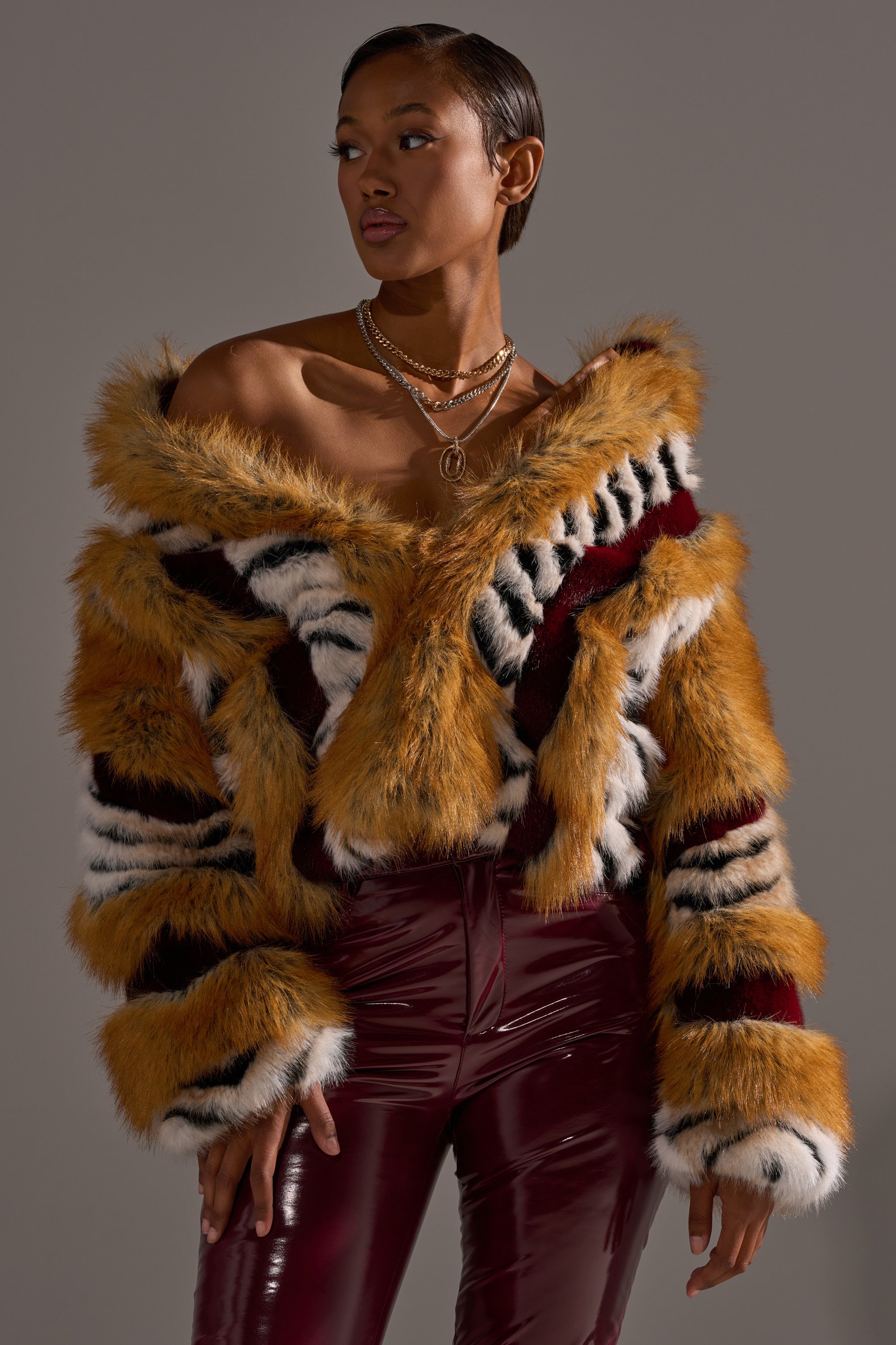 A woman models the OLYMPIA PANELED FAUX FUR coat with orange, black, white, and red panels, styled with shiny burgundy pants and layered gold necklaces against a neutral backdrop.
