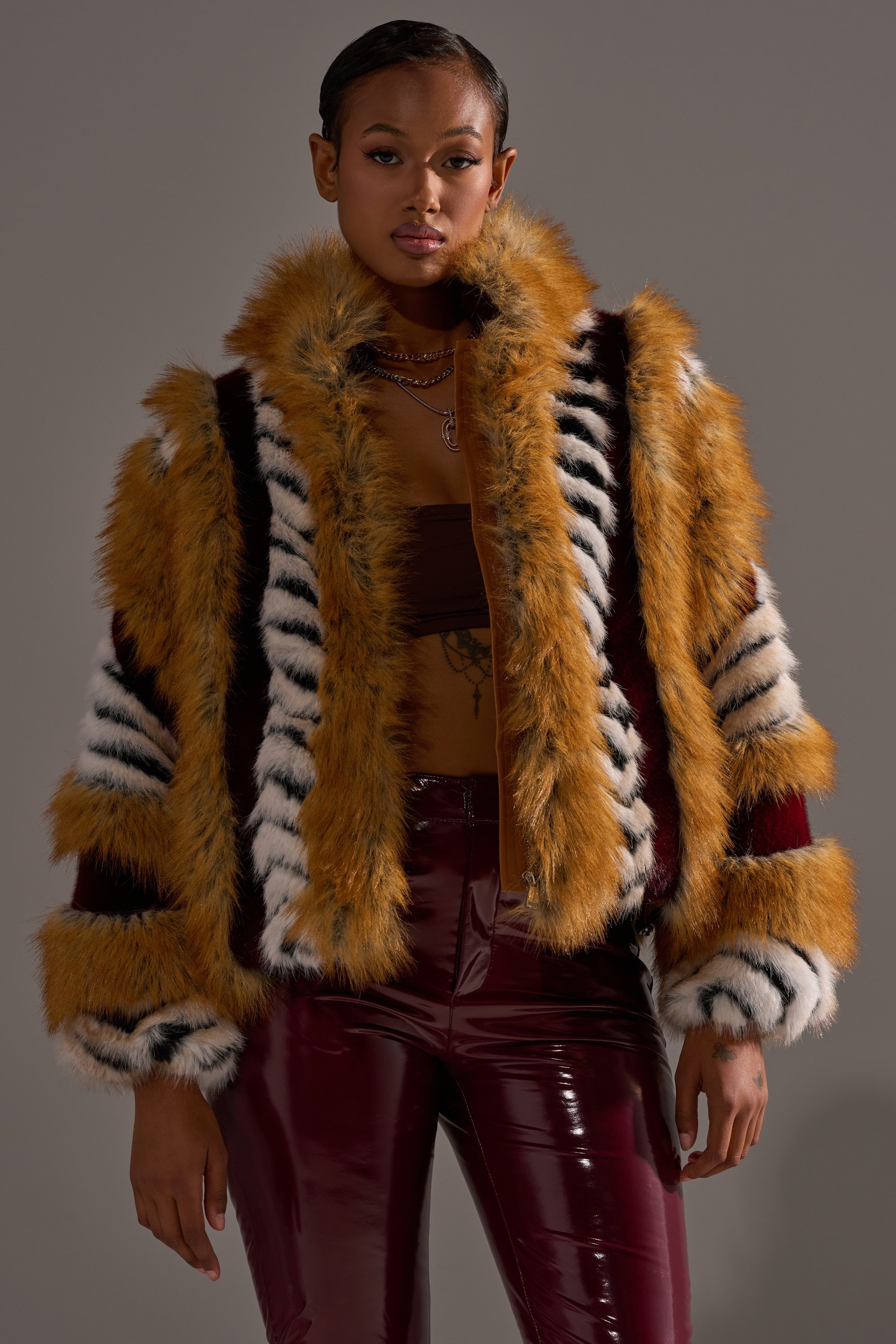 A woman wears the OLYMPIA PANELED FAUX FUR jacket, featuring orange, black, and white stripes, over a brown crop top and shiny burgundy pants against a plain gray background.