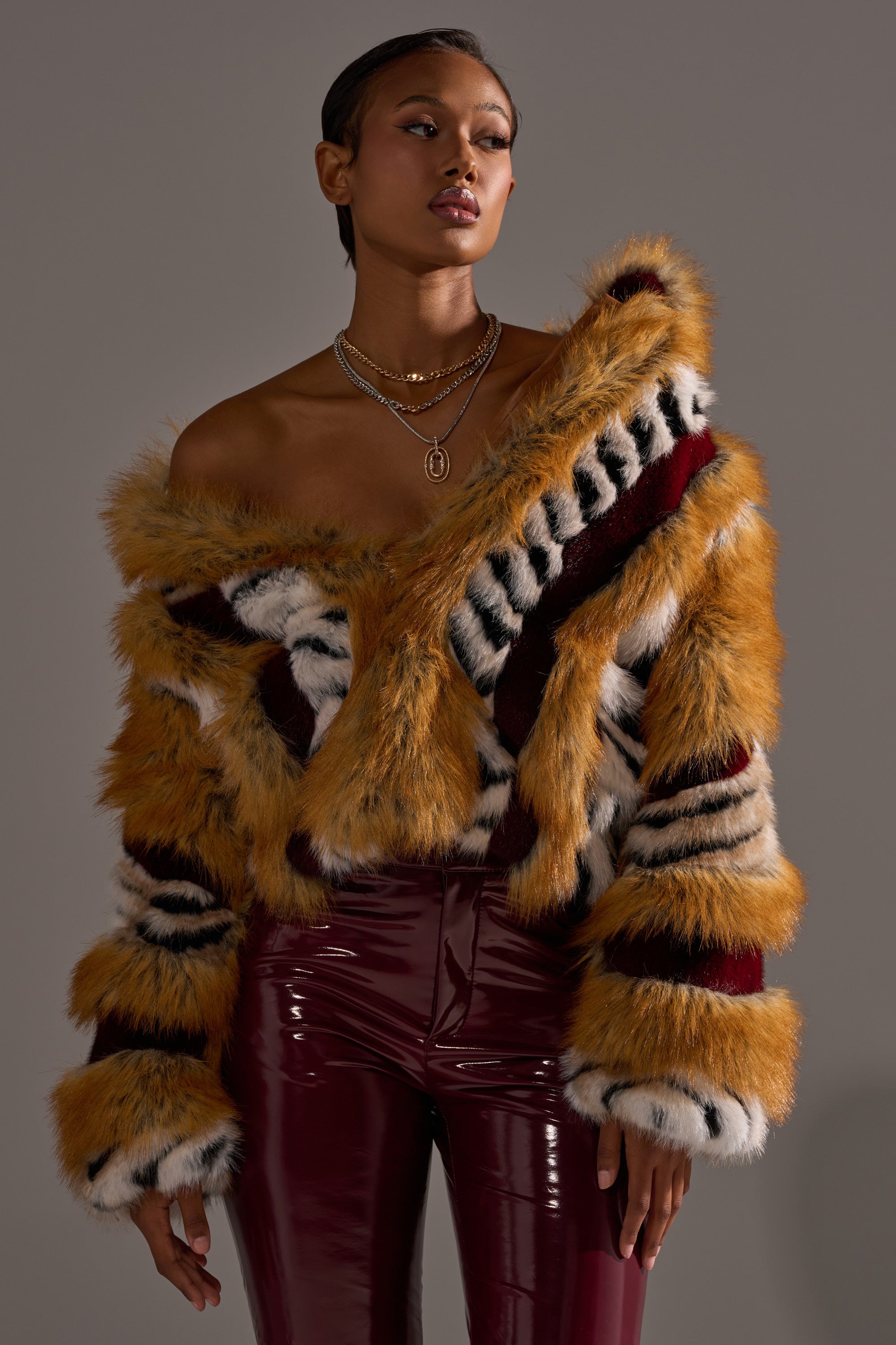 A woman poses in the OLYMPIA PANELED FAUX FUR jacket, featuring bold gold, black, white, and burgundy panels, styled off-the-shoulder with shiny burgundy vinyl pants and layered gold necklaces against a neutral background.