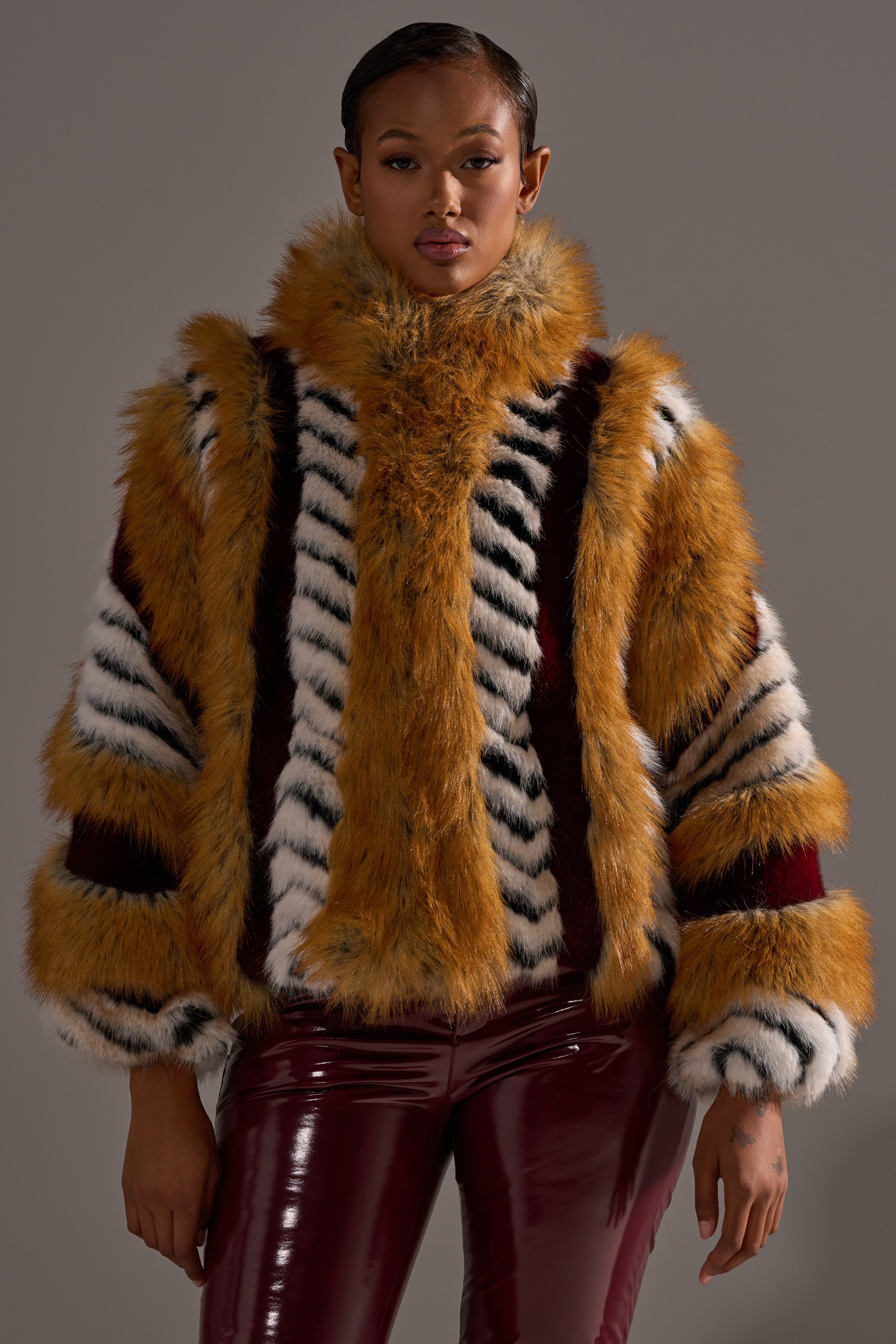 Wearing the OLYMPIA PANELED FAUX FUR, featuring bold orange, white, and black stripes, and shiny burgundy pants, a person stands confidently against a plain gray background.
