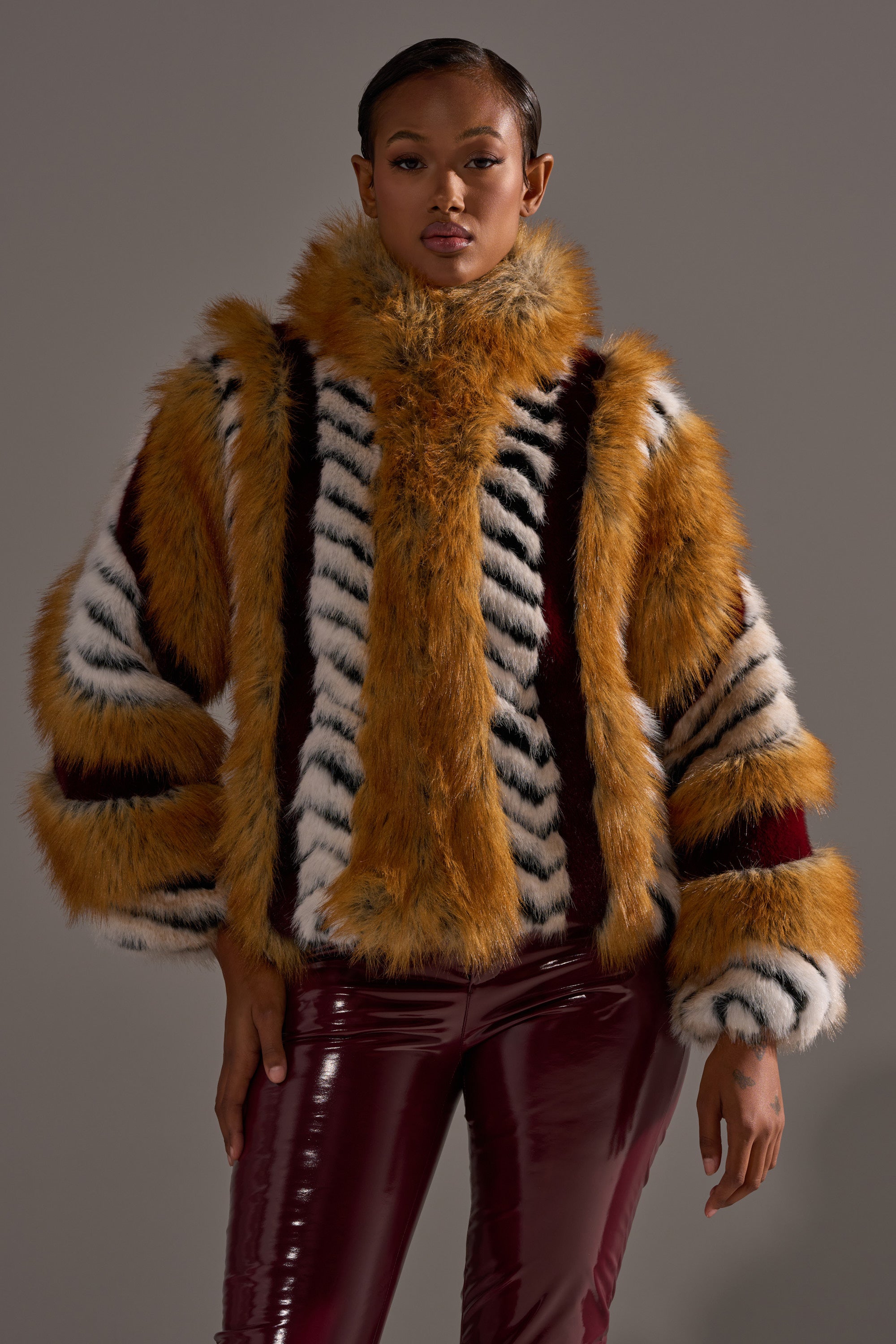 A woman faces the camera confidently against a gray background, wearing the OLYMPIA PANELED FAUX FUR jacket with orange, white, and black stripes, paired with glossy maroon pants.
