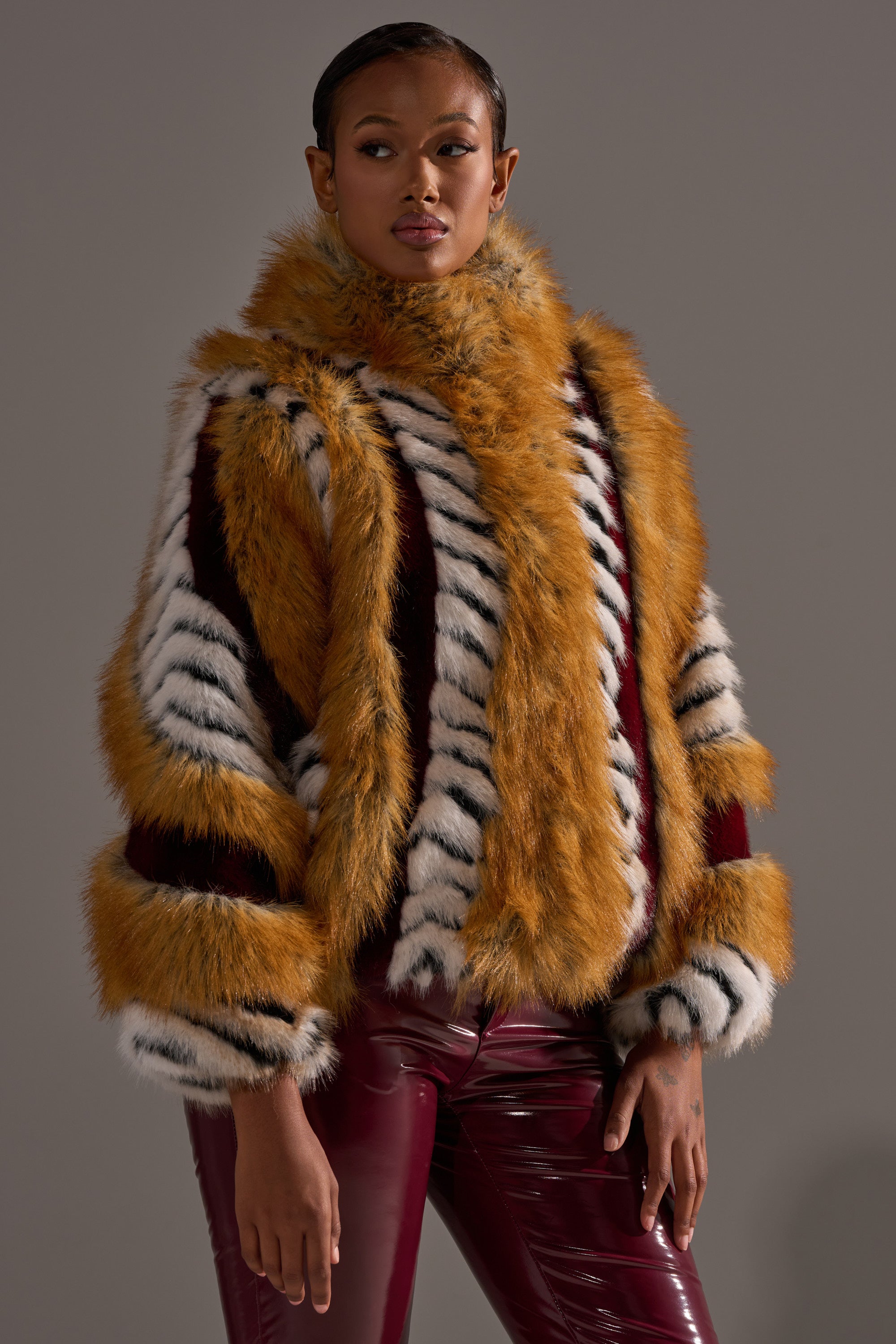 A woman wearing the OLYMPIA PANELED FAUX FUR coat with gold, white, black, and red stripes pairs it with shiny burgundy pants, standing against a plain gray background.
