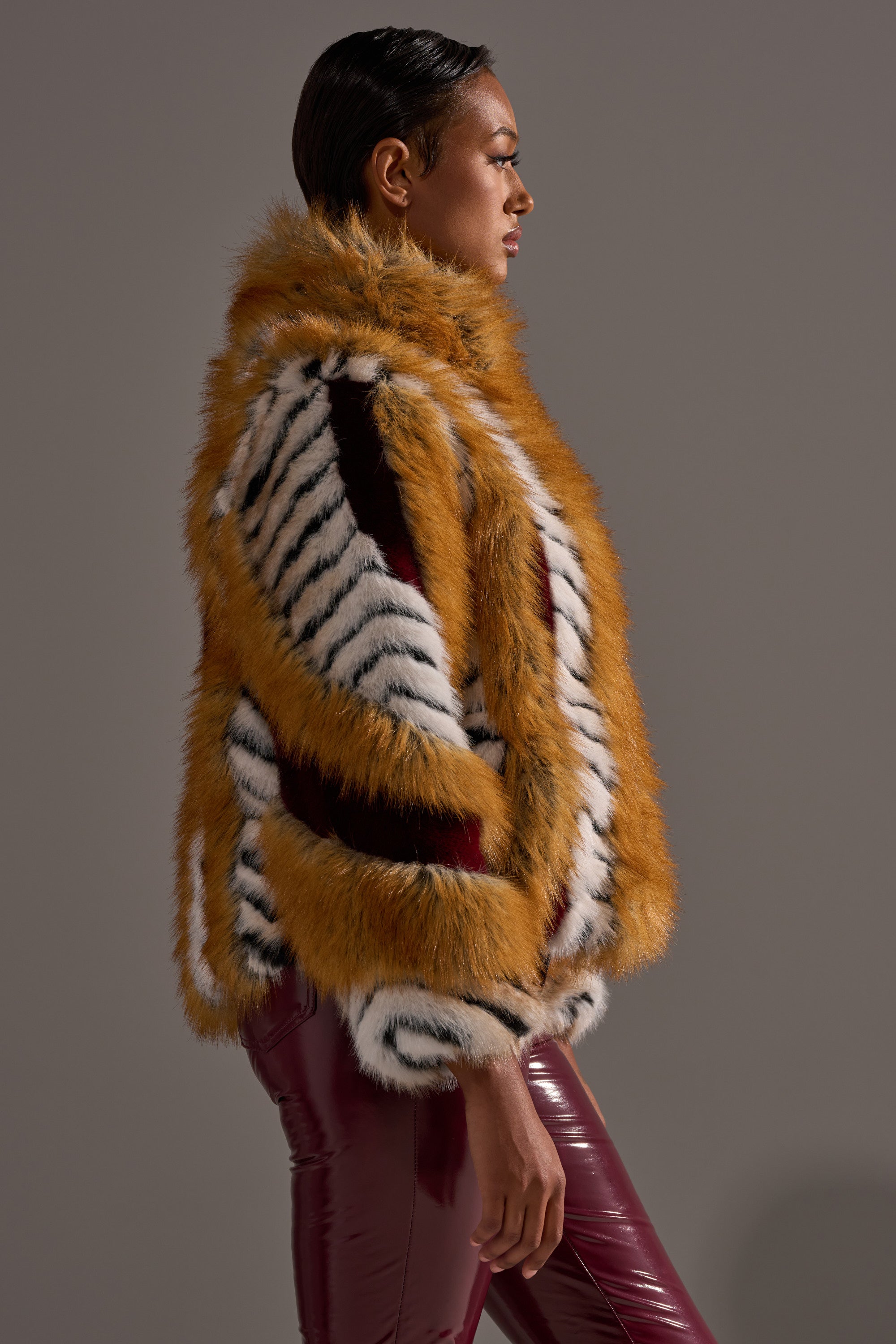 A person in profile models the OLYMPIA PANELED FAUX FUR coat—oversized with orange, white, and black stripes—paired with shiny burgundy pants against a plain gray background.