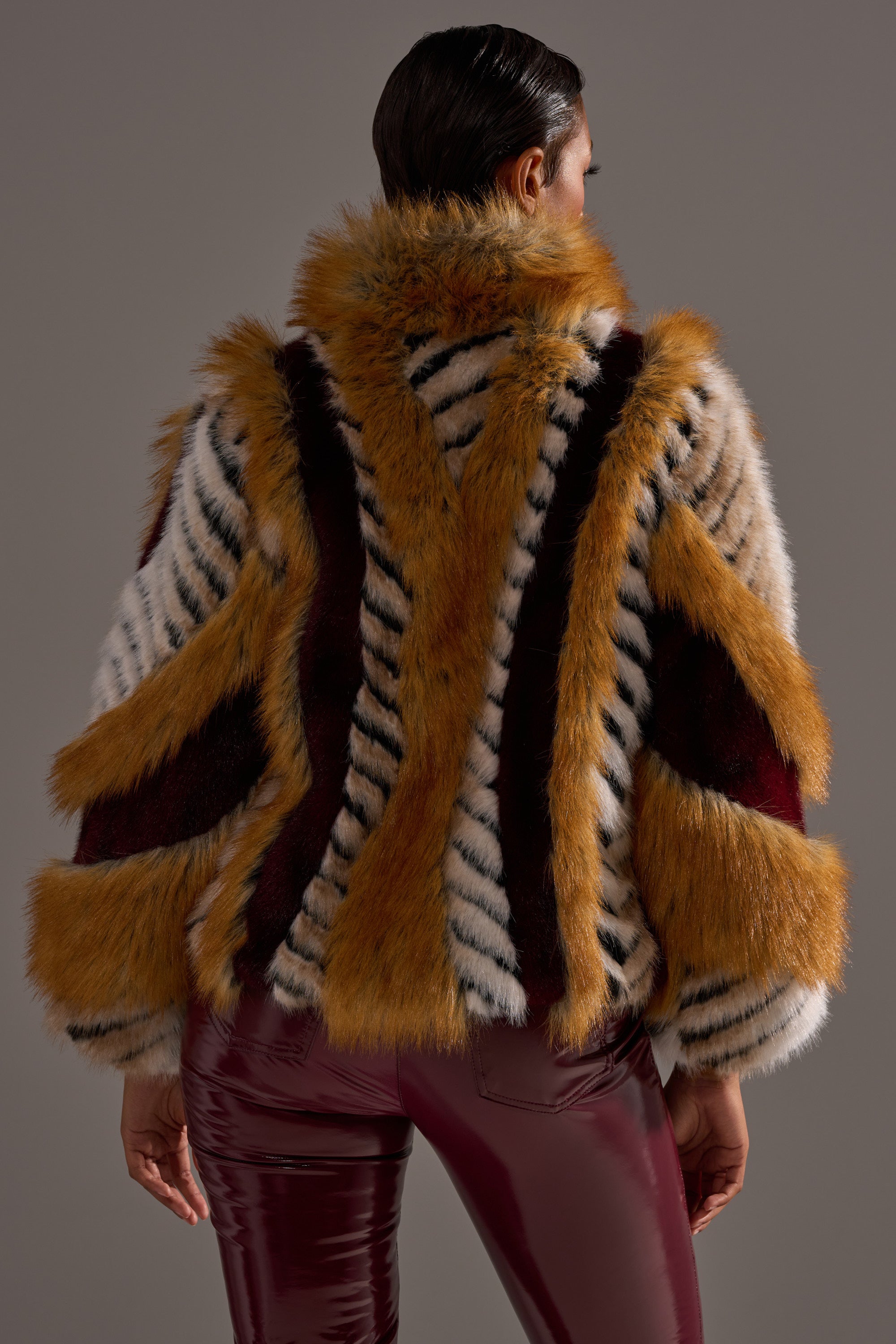 A person faces away, wearing the OLYMPIA PANELED FAUX FUR coat with orange, black, white, and burgundy stripes over glossy burgundy pants. The bold textured pattern and oversized fit create a striking look with slicked-back hair for added edge.