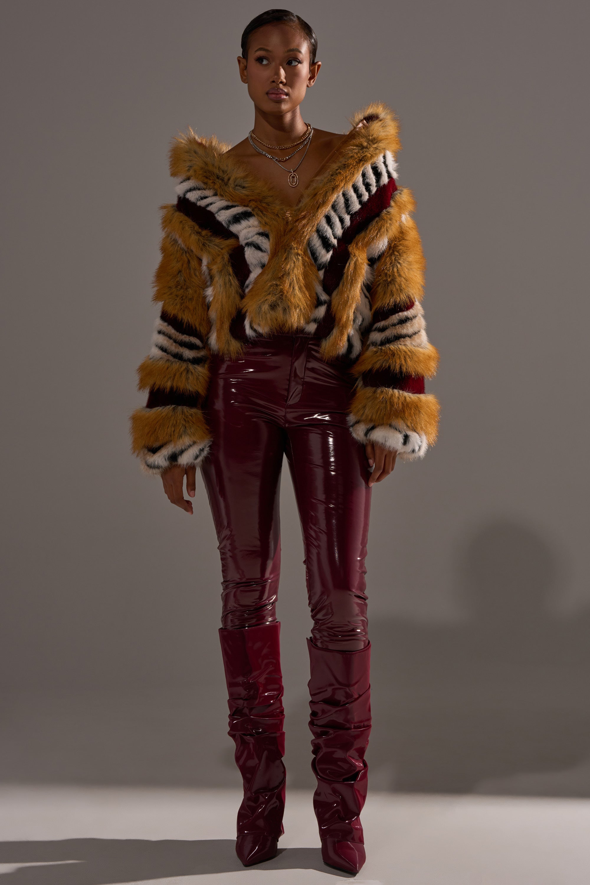 A woman poses against a gray background in the OLYMPIA PANELED FAUX FUR coat with zigzag patterns, paired with shiny burgundy vinyl pants, matching high-heeled boots, and layered gold necklaces.