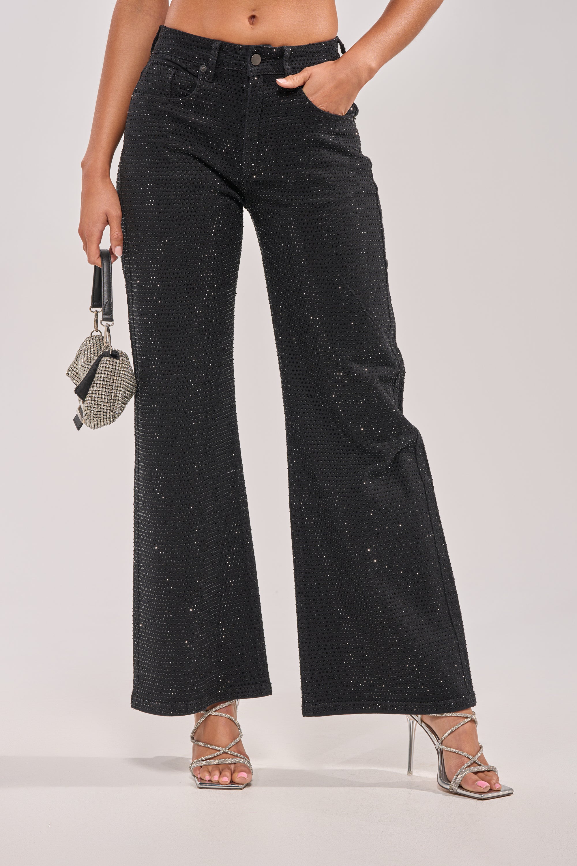 A person wearing SHINING BRIGHT RHINESTONE DENIM high-waisted black sparkle pants, silver strappy heels, and holding a small rhinestone-studded bag against a plain background. 1