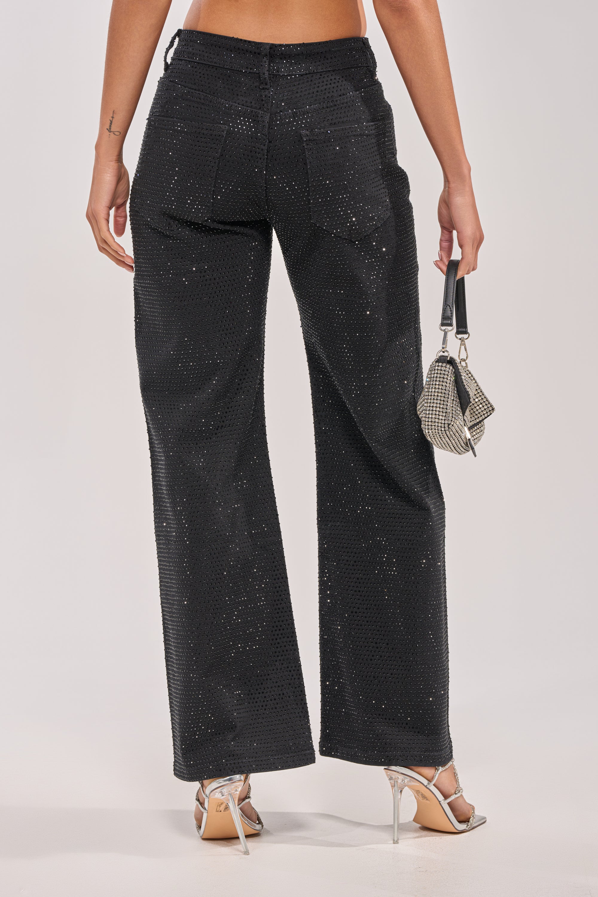 A person in SHINING BRIGHT RHINESTONE DENIM wide-leg jeans and silver high heels holds a small textured handbag, seen from behind against a plain background. 3