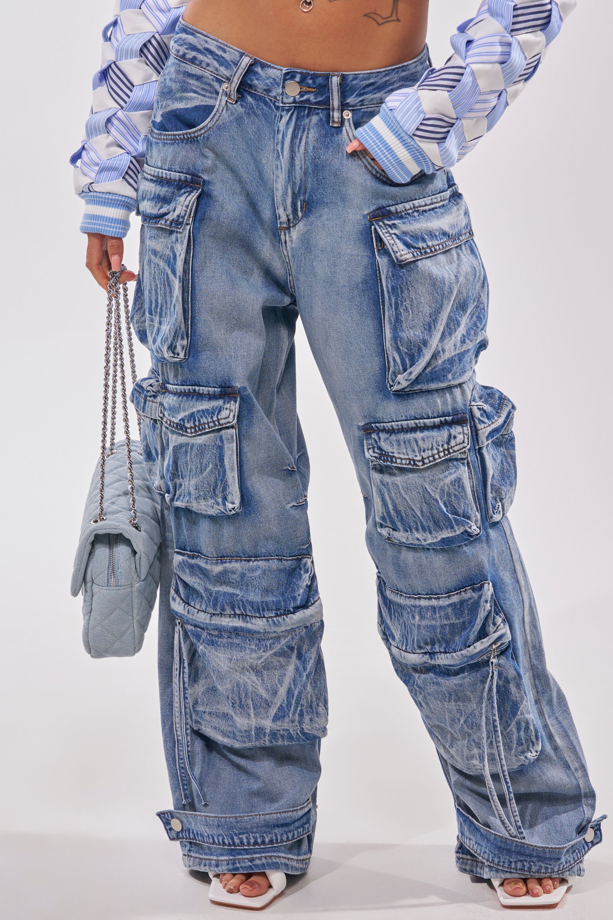 Someone in EASY ENOUGH CARGO DENIM—loose-fitting, light blue cargo jeans with large pockets and white streaks—styled with a blue quilted handbag, white sandals, and a patterned blue-and-white long sleeve top for a trendy streetstyle look. 0