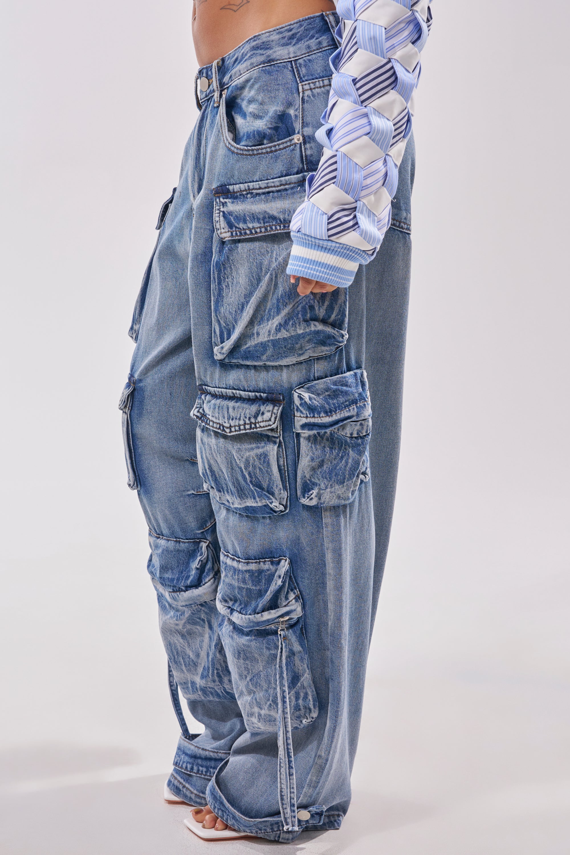 Wearing the EASY ENOUGH CARGO DENIM—light-wash, loose cargo jeans with multiple big pockets—paired with a geometric long-sleeve shirt. Cropped at the torso, bare feet visible, capturing relaxed streetstyle vibes. 2
