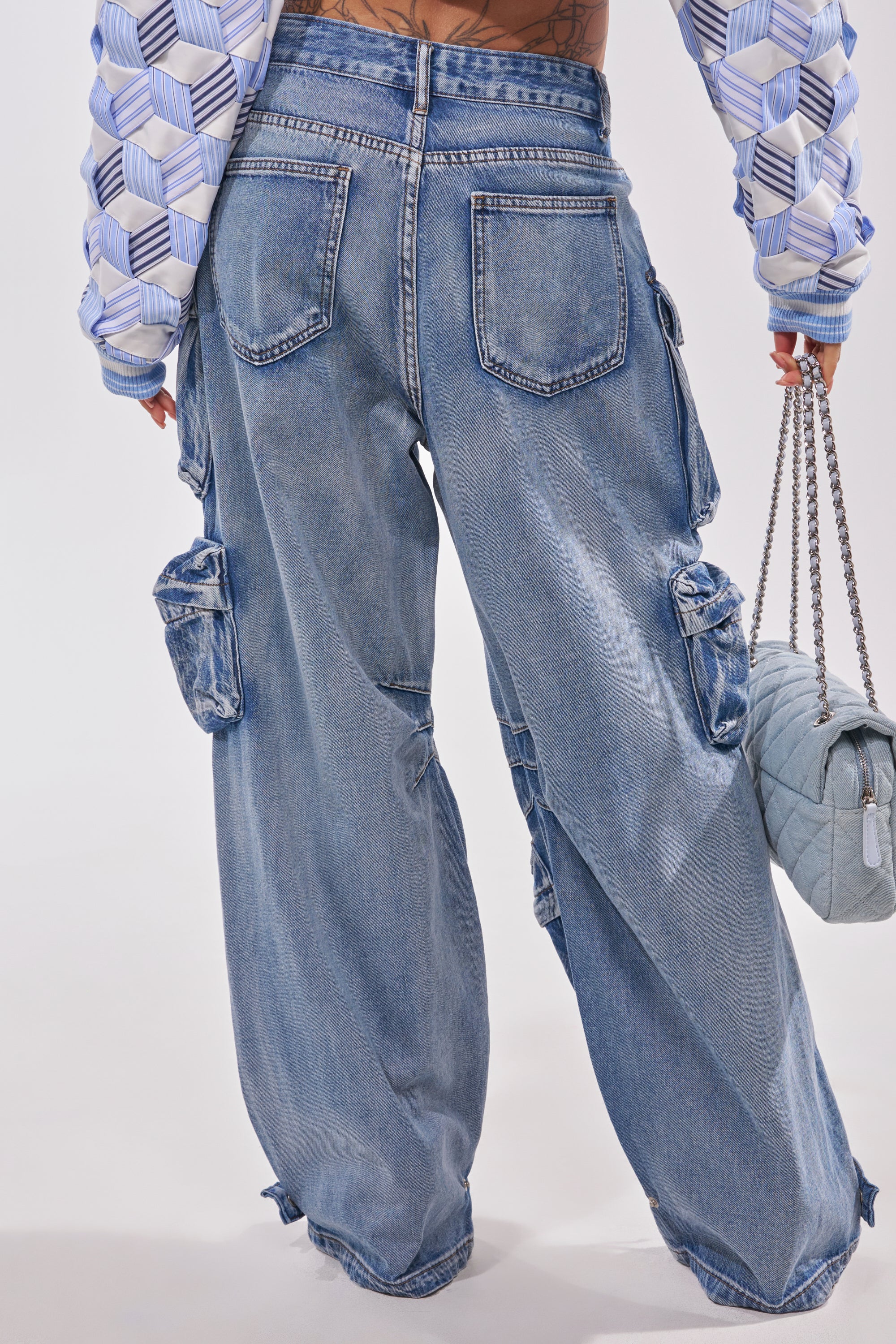 A person wears the EASY ENOUGH CARGO DENIM—loose, light blue streetstyle pants with multiple pockets—paired with a patterned, geometric long-sleeve top and a quilted blue chain-strap purse. 3