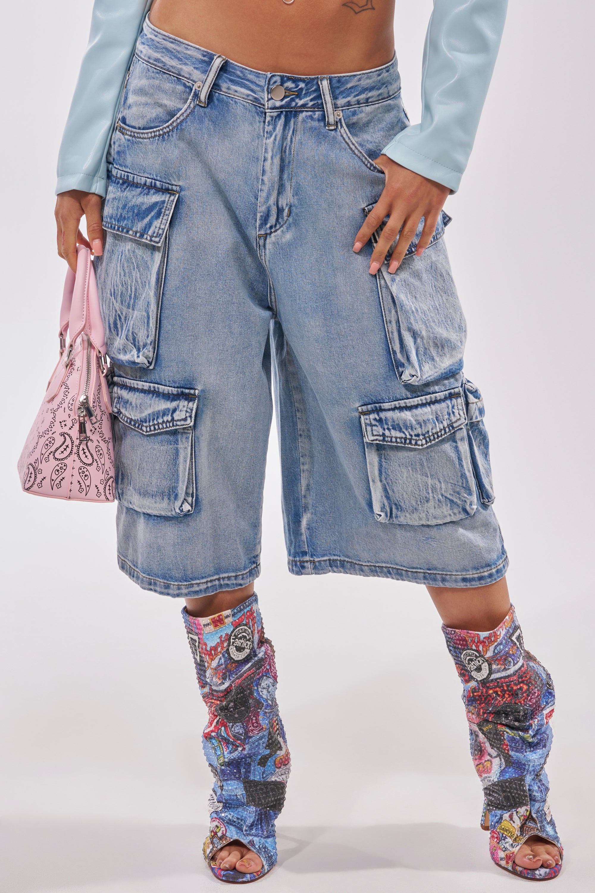 Someone shows off festival style in knee-length, light-wash TOO MUCH FUN CARGO BERMUDA SHORTS with big pockets, paired with a light blue top, colorful patterned boots, and a pink paisley handbag. 1