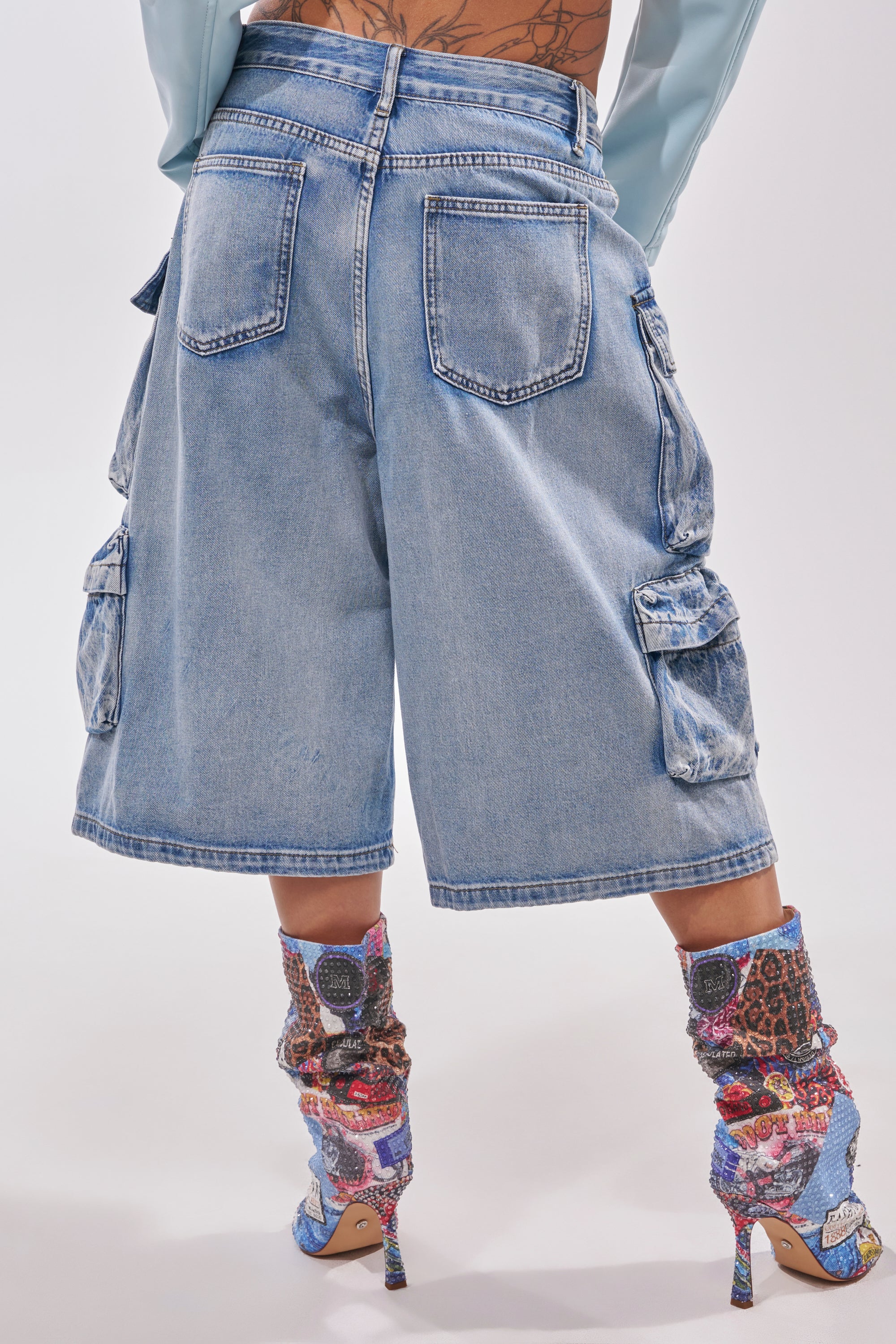 A person styles the TOO MUCH FUN CARGO BERMUDA SHORT with bold patterned high-heeled boots and a long-sleeve top, showing off large pockets and back tattoos for a striking festival-inspired streetwear look. 3