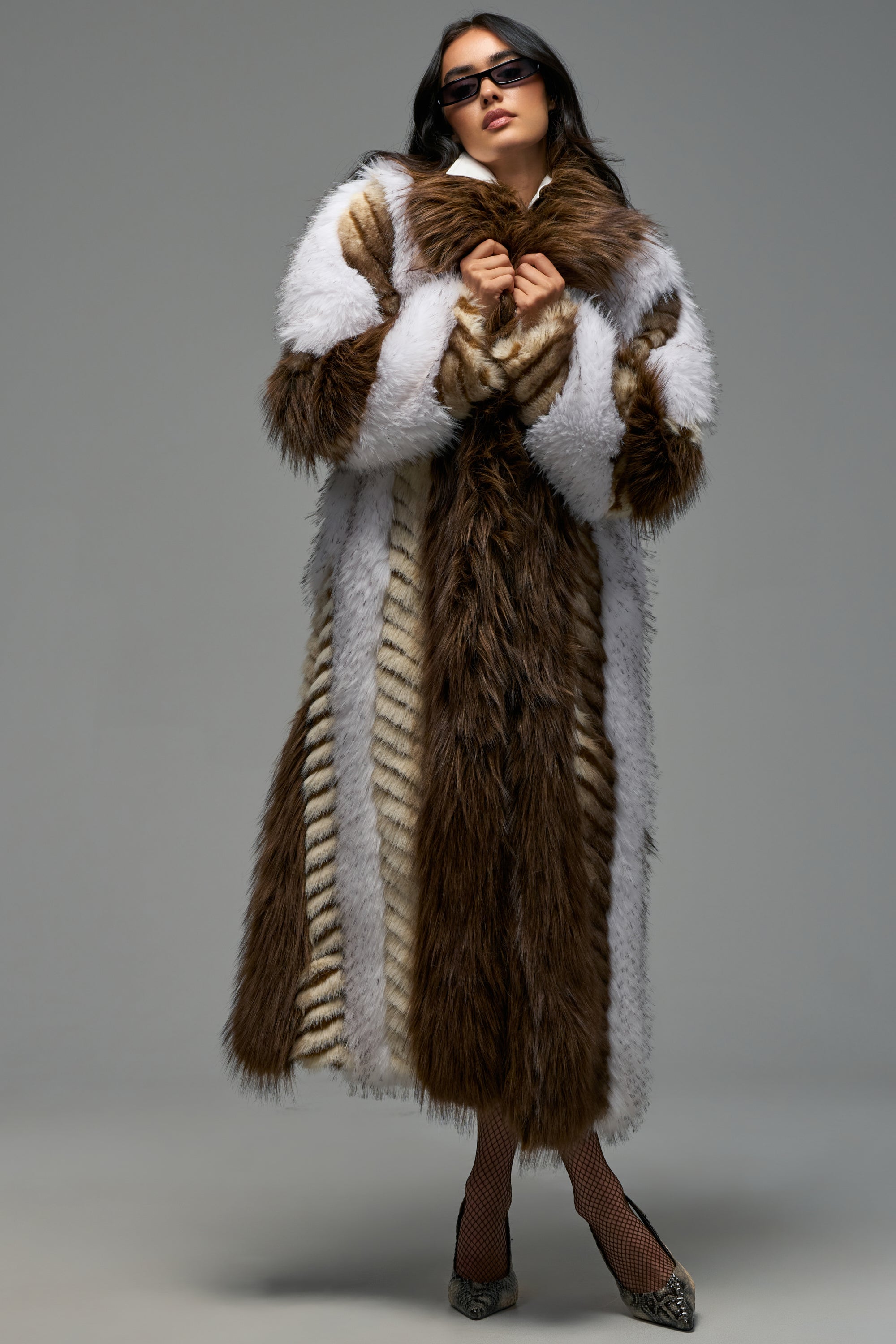 A woman wearing sunglasses poses against a plain gray background, showcasing the OMEGA PATCHWORK FAUX FUR—a luxurious statement winter coat featuring bold brown, white, and striped patterns. She has her hands near her collar and completes the look with fishnet stockings and heels.