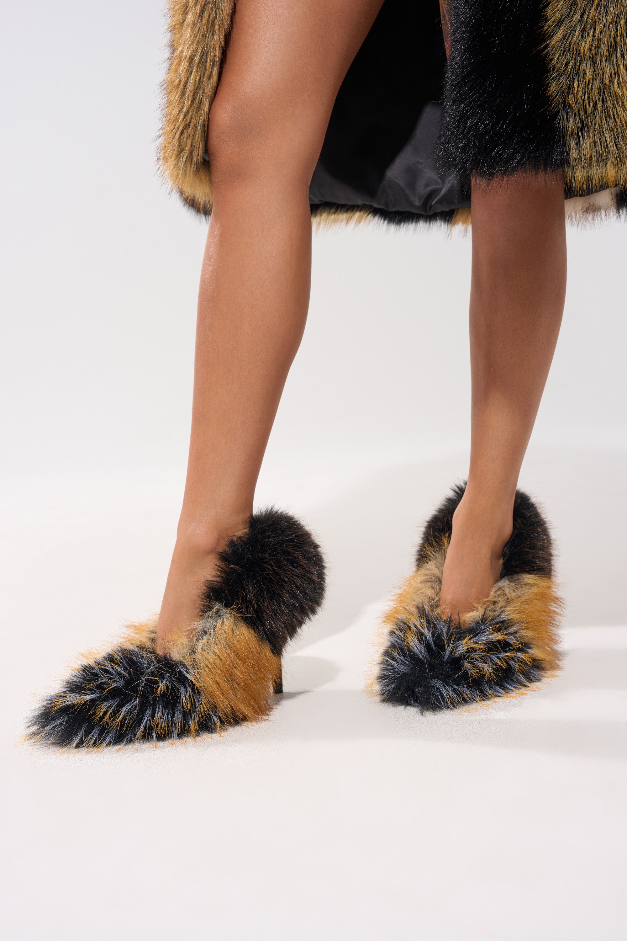 Only the legs and striking AZALEA WANG OMICRON BLACK AND TAN FUR STILETTO PUMPS are visible as someone stands on a light surface, wearing a matching long brown and black faux fur coat.