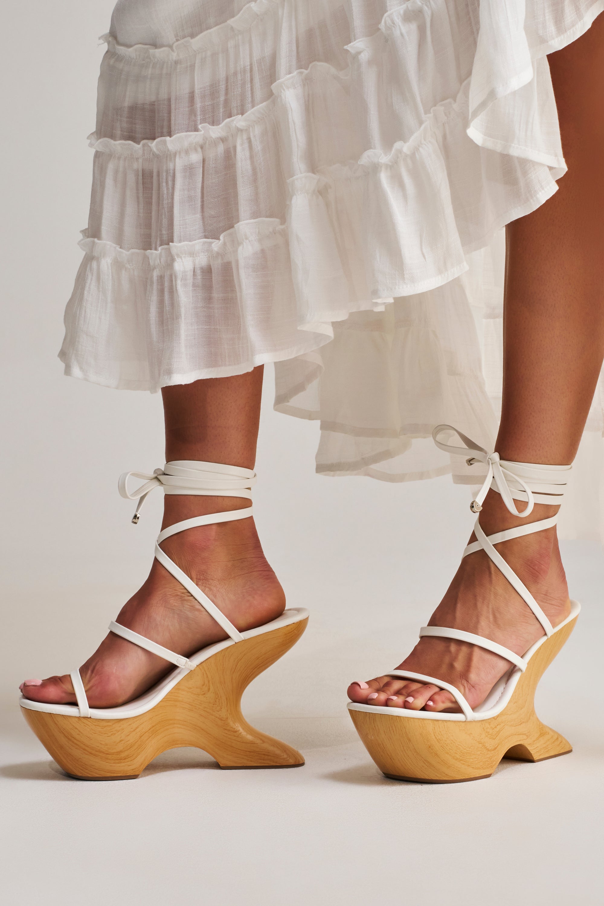 A person wearing a white, ruffled, ankle-length skirt and AZALEA WANG ONNI WHITE STRAPPY UPPER WOOD WEDGE sandals with uniquely curved wooden soles stands on a light-colored surface.
