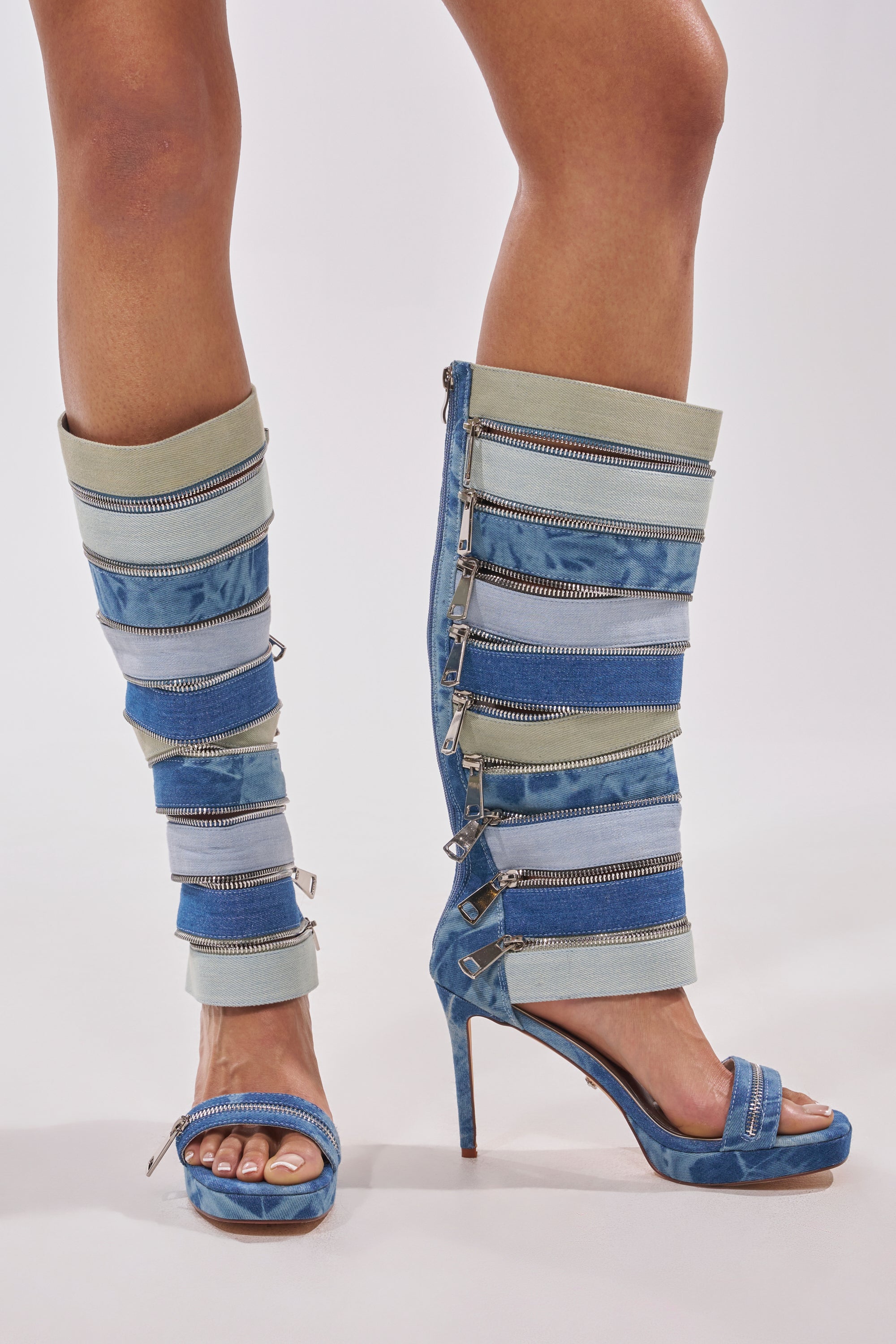 A person wears the AKIRA GOLD ONREPEAT DENIM EMBELLISHED PLATFORM STILETTO SANDAL BOOT, featuring knee-high denim with layered strips and multiple zippers, styled with blue open-toe heels against a plain white background.