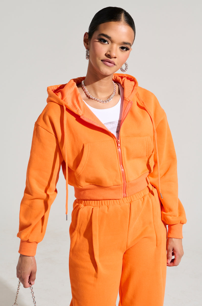 A person wearing the ORANGE CROP ZIP UP SWEATSHIRT paired with matching pants exudes confidence against a plain background. The cotton blend outfit is enhanced with silver jewelry, featuring a chain necklace and hoop earrings.