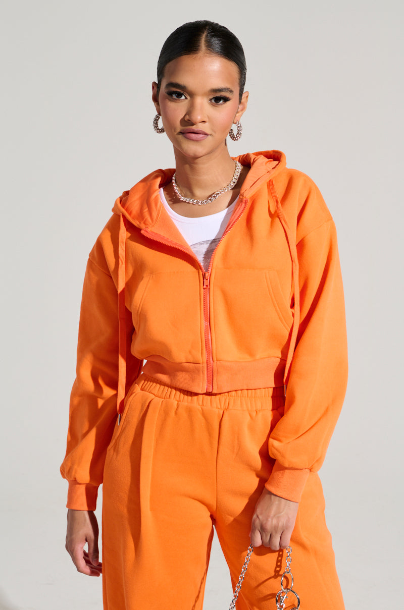 A person clad in the ORANGE CROP ZIP UP SWEATSHIRT with a hood stands confidently against a neutral background. With their short hair pulled back, they accessorize with large hoop earrings and a chain necklace. Holding a chain in their right hand, they exude effortless style.