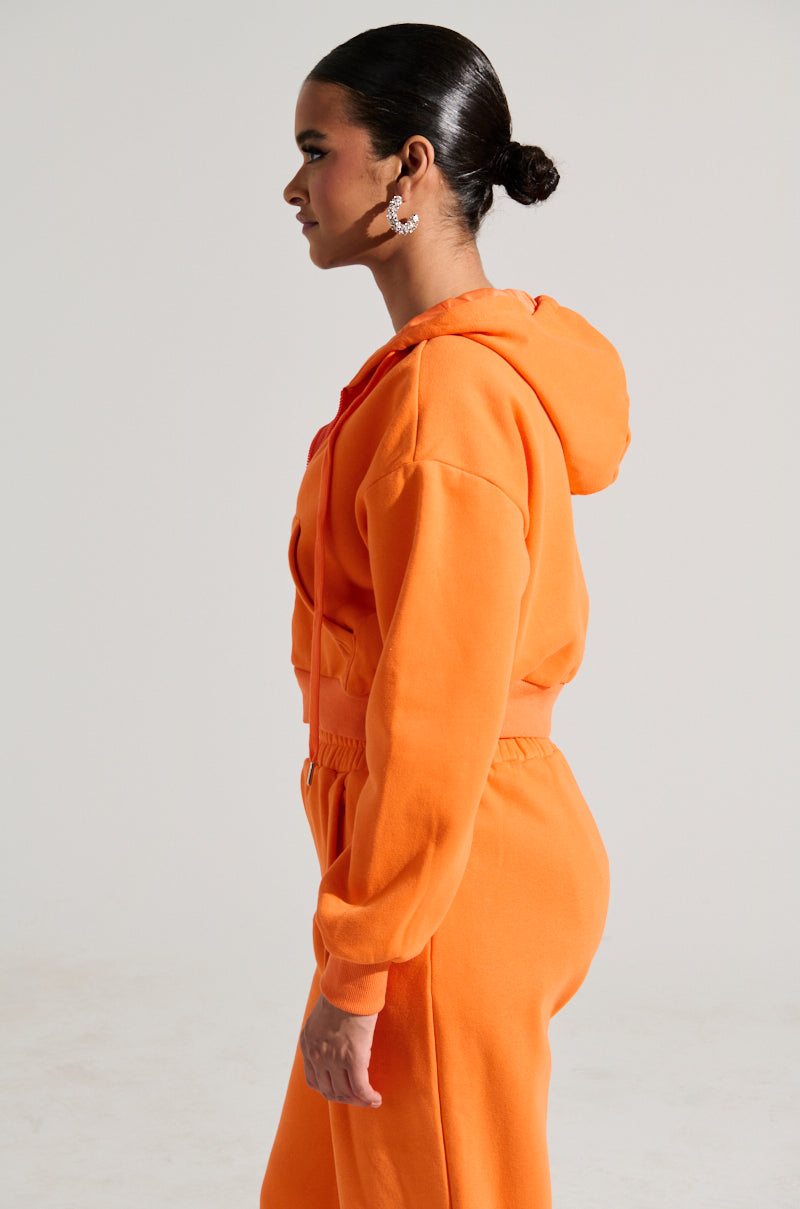 A person in profile view is wearing the ORANGE CROP ZIP UP SWEATSHIRT along with matching sweatpants. Made from a comfortable cotton blend, the outfit perfectly complements their dark hair styled in a bun as they stand against a plain background, showcasing casual elegance with a touch of sporty flair.