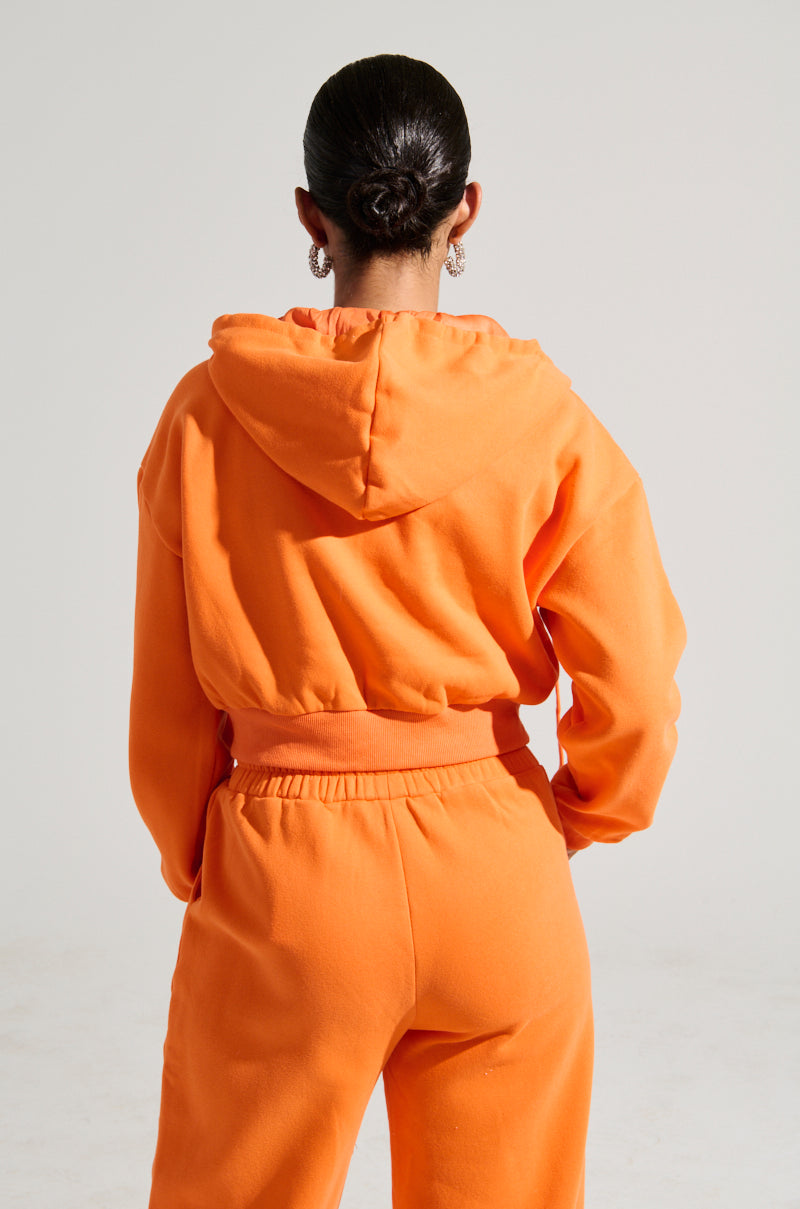 Wearing the ORANGE CROP ZIP UP SWEATSHIRT, a person with their hair tied in a bun and matching sweatpants made from a semi-stretchy cotton blend is seen facing away from the camera against a neutral background.