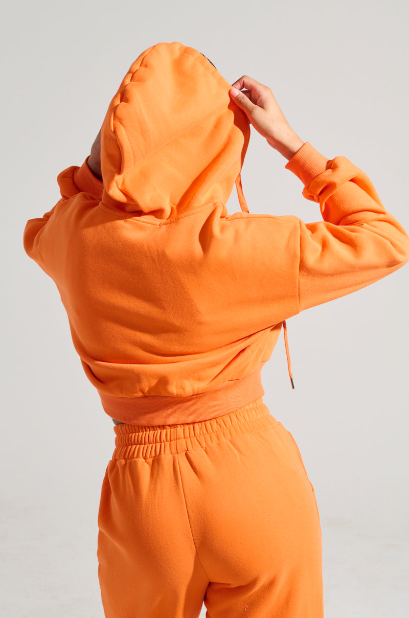 A person in an ORANGE CROP ZIP UP SWEATSHIRT and cotton blend sweatpants, seen from the back, adjusts the hood.