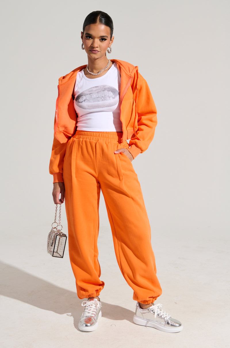Dressed confidently in the ORANGE CROP ZIP UP SWEATSHIRT over a white graphic T-shirt and matching ribbed knit tracksuit, a person holds a small, glossy purse and dons white sneakers. The plain background highlights their sporty and stylish ensemble.